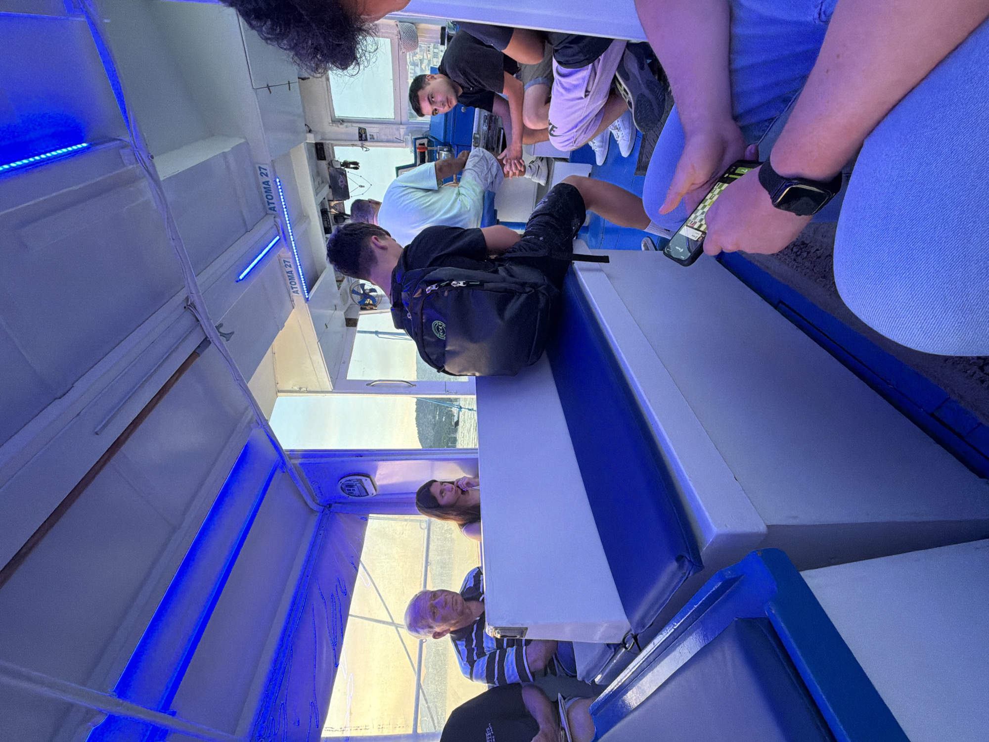 This image shows the interior of a boat cabin, likely a passenger ferry or a tour boat, with several people onboard. Bright blue LED strip lights illuminate the white ceiling and walls, creating a distinctive ambiance. Through the windows, warm, golden sunlight indicates late afternoon or early evening, revealing distant hills or land, aligning with the coastal environment of Poros, Greece.
In the foreground on the right, a person wearing light blue jeans holds a smartphone that displays a game, possibly chess, and wears a smartwatch. Towards the center, a young man with a black backpack is seated with his back to the camera. Another young man, dressed in a black t-shirt and shorts, is seen standing and looking towards the camera. Behind him, an older man in a light shirt is seated with his back to the viewer. Further into the cabin on the left, an older man in a striped shirt is seated, looking towards the camera, while a woman with long dark hair is visible behind him.
The boat's interior features white and blue seating and surfaces, and a small fan is visible on the left side. The text "ATOMA 27" is inscribed twice on the white wall, suggesting capacity or section numbering, with "ATOMA" being the Greek word for individuals. The scene portrays a casual journey or leisure activity, with passengers relaxing and engaging with their personal devices or observing their surroundings.