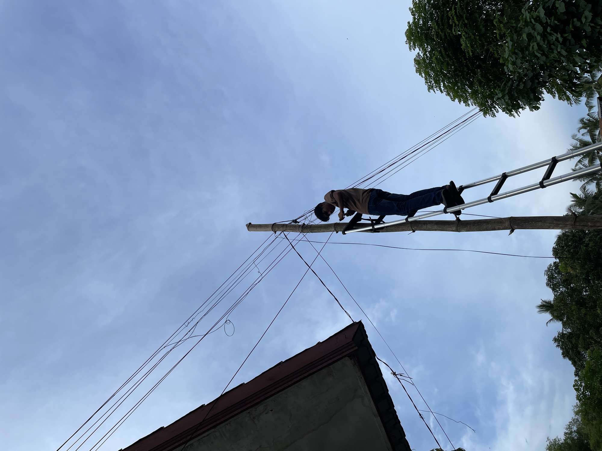 A low-angle outdoor photograph captures a person engaged in work on an overhead utility setup. A man, dressed in a brown shirt and blue jeans, is positioned horizontally on a metal ladder. The ladder is propped against a thick wooden pole, from which numerous black wires and cables extend in various directions. The man appears to be inspecting or working on these connections.
The sky, a soft blue with wispy clouds, dominates the upper portion of the frame, indicating daytime. Lush green foliage from tropical trees, including what appears to be a palm, frames the upper right and right side of the image. In the lower left, the concrete and terracotta-tiled roof of a building is visible, with more wires leading towards it. The overall setting suggests a semi-rural or residential area in Grong-Grong Gogo, Indonesia. The worker's precarious position on the ladder highlights the demanding nature of the task. No visible text is present in the image.