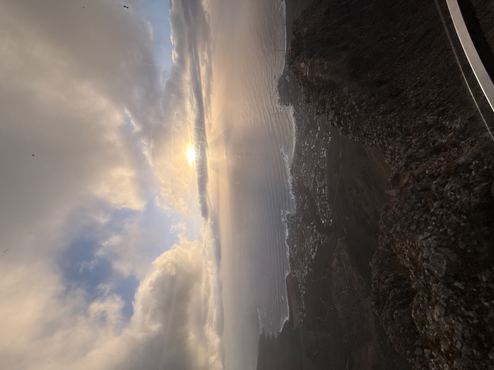 The image shows an aerial view of the coastline and cityscape, most likely taken from the upper reaches of Table Mountain in Cape Town, South Africa, at dusk. The setting sun, obscured by clouds, casts a golden light on the ocean and the landscape. The coastline is rugged, with waves crashing against the shore. The city sprawls inland from the coastline, with buildings and roads visible in the distance. There are no discernible people or animals.