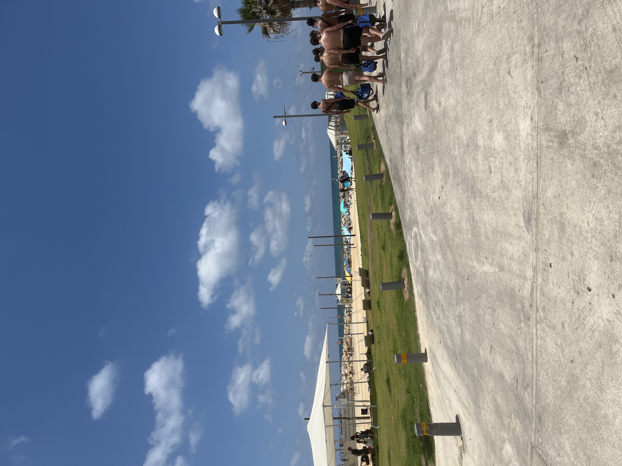 The image depicts a sunny day at a beach in Tel Aviv, Israel, likely on the promenade adjacent to the sand. In the foreground, a concrete pathway is visible, with several bollards lining its edge. A group of young men, mostly shirtless and wearing shorts, are walking away from the camera along this pathway, some carrying blue duffel bags. To the left, a grassy area is bordered by the pathway, with more bollards. Beyond the grass, a section of the beach is visible, featuring a large white canopy providing shade for people sitting or lying down. Further towards the sea, beachgoers can be seen relaxing on the sand and near the water. The ocean stretches to the horizon under a bright blue sky dotted with scattered white clouds. A palm tree is visible on the right side of the image, near a lamppost. The overall scene conveys a relaxed atmosphere of a summer day at the beach.