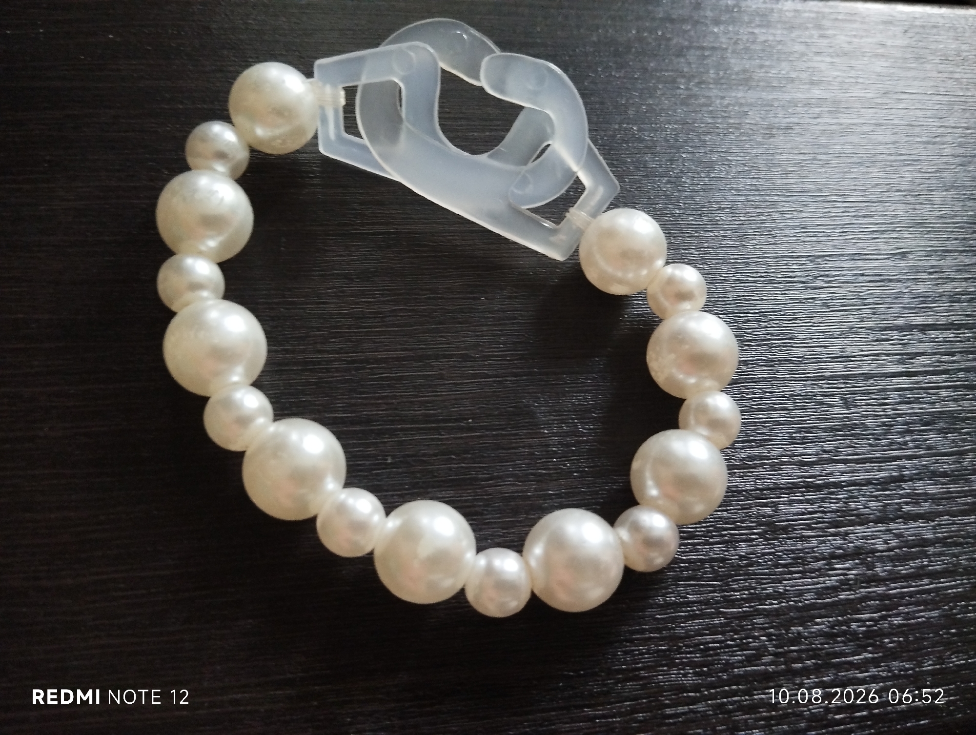An overhead shot shows a pearl bracelet with a clear plastic clasp, laid out on a dark, textured wooden surface. The bracelet is composed of various sizes of white, iridescent pearls. The clasp is a distinctive interlocking design, appearing to be made of a matte, translucent plastic. The background is a dark, horizontal-grained wood, suggesting a table or desk. There are no people visible in the image. The bottom left corner displays the text "REDMI NOTE 12", and the bottom right corner shows a date and time stamp: "10.08.2026 06:52".