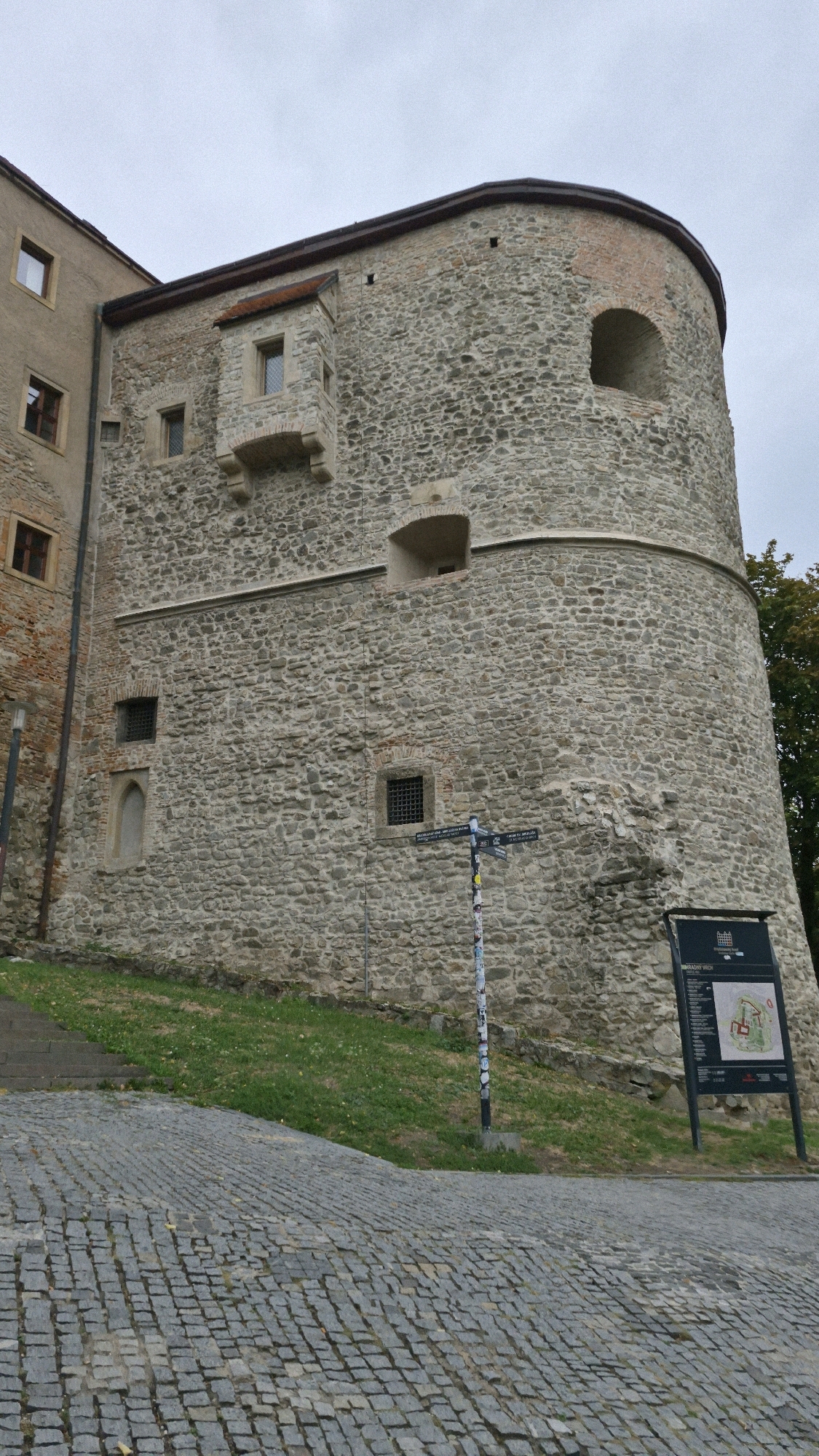 The image shows a view of Bratislava Castle in Bratislava, Slovakia. The castle is a historic landmark that overlooks the city. In the foreground, there is a cobblestone path leading up to the castle. The castle itself is made of stone and has a large, round tower. There is a sign with information about the castle and its grounds. The sky is overcast, and there are some trees in the background.