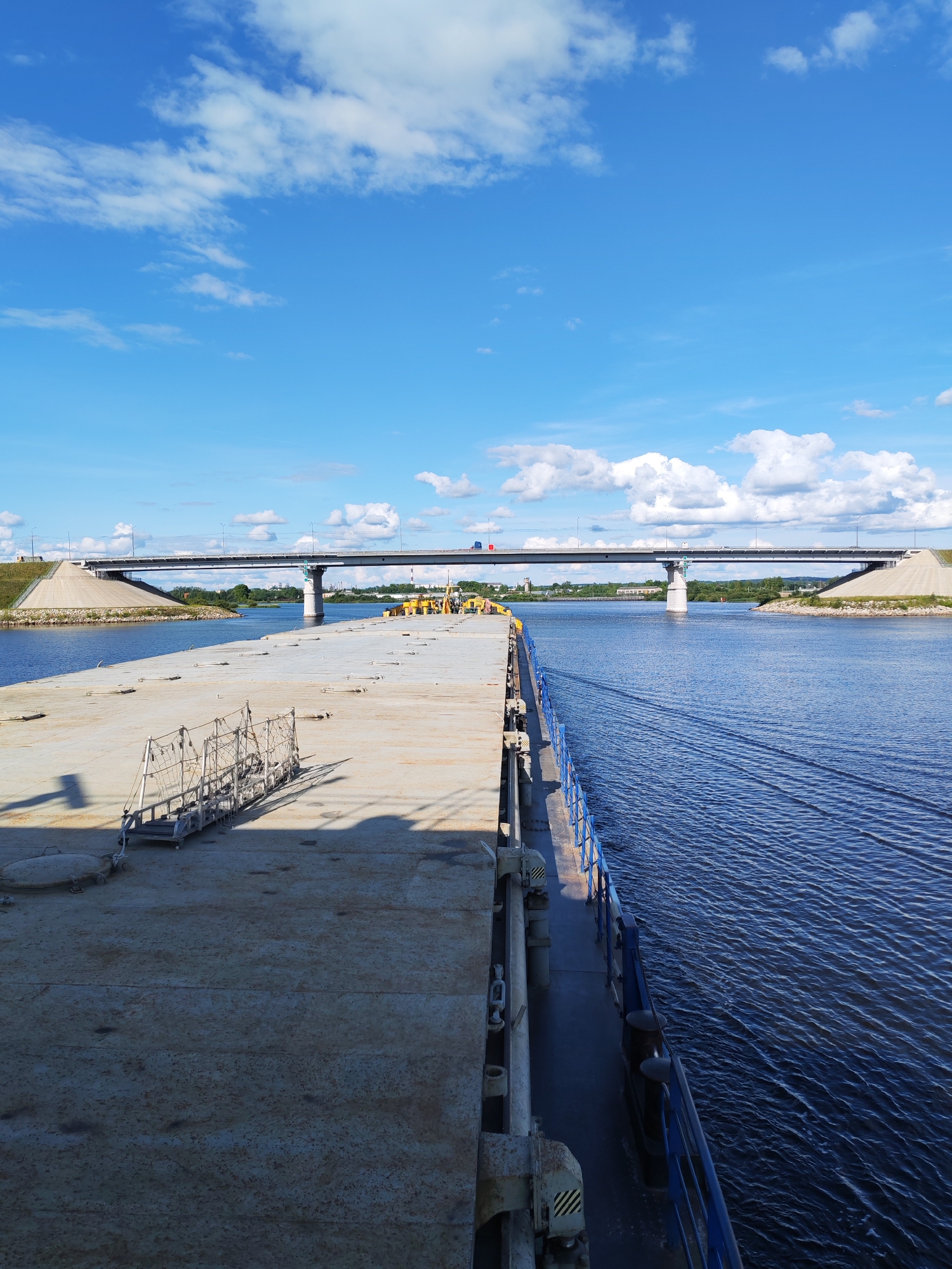 An exterior, wide-angle, eye-level shot captures a barge navigating a wide, calm river under a bright blue sky filled with scattered white clouds. The barge occupies the left and foreground of the image, its flat, metallic deck extending towards the horizon. A blue metal railing runs along the edge of the barge. A narrow gangway with three levels of steps and handrails is positioned on the barge's deck, casting a long shadow. In the mid-ground, a modern concrete and steel bridge spans the river, with a single lane of traffic visible on its surface. The riverbanks are lined with grassy slopes and some scattered trees. The scene is set in or near the city of Sheksna, Russia, likely during daylight hours with clear weather.