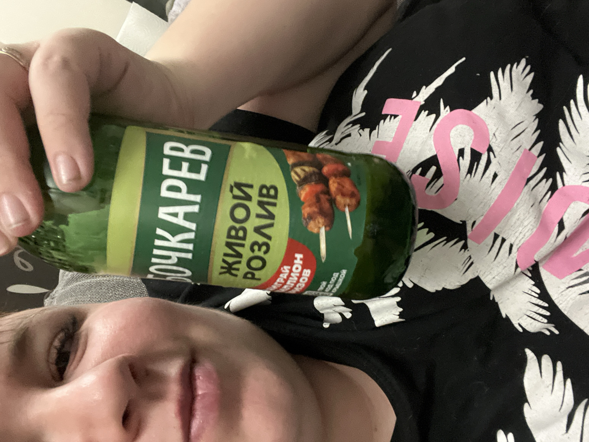 A person is holding a bottle of Bochkarev beer, Живой Розлив (Zhivoy Rozliv, Live Bottling), in a selfie. The bottle's label shows a picture of shashlik (grilled meat skewers). The person is wearing a black shirt with white palm tree designs and pink lettering. The location is likely Chaikovsky, Russia, given Bochkarev beer is a Russian brand.