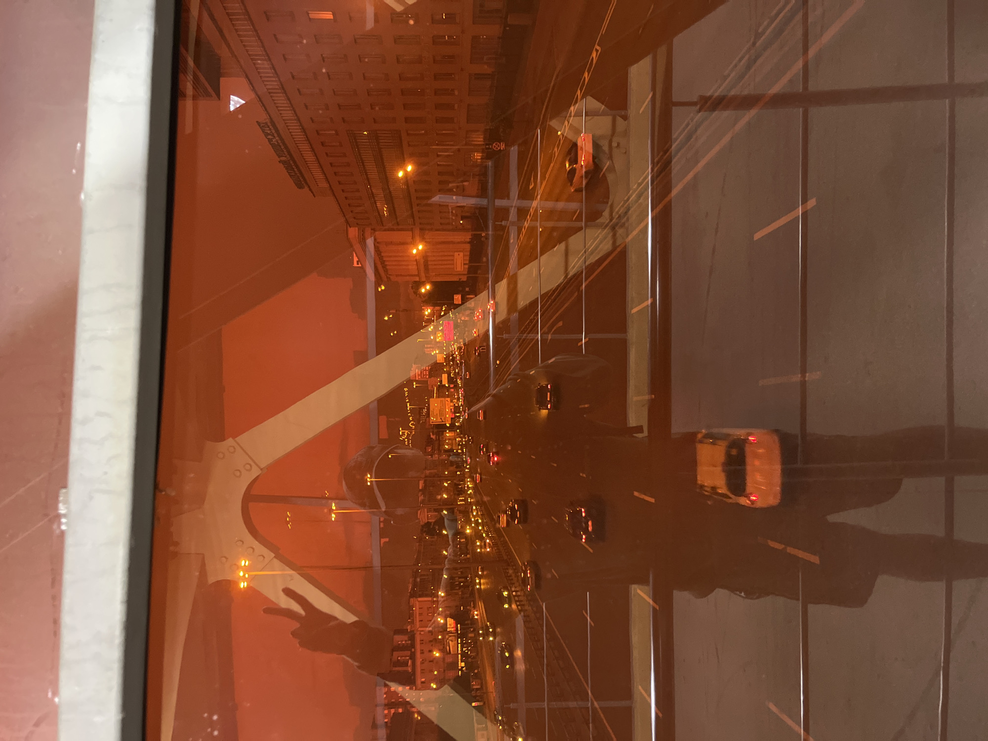 The image depicts a reflection on an orange-tinted window, offering a view of a cityscape at dusk or night. A person's silhouette is visible, holding up a hand in a peace sign. The city scene includes a multi-lane highway with traffic, illuminated by streetlights and the headlights of vehicles. Buildings line the road, and the overall atmosphere is urban and active. Based on the inscription on the building, "Московский Государственный Университет", the location is identified as Moscow State University, which is in Moscow, Russia, not Khimki.