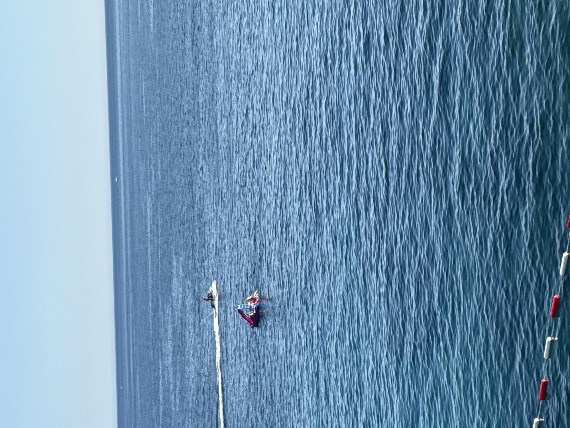 In the open sea off the coast of Kamenovo, Montenegro, two distinct watercraft are visible. In the distance, a single person navigates a jet ski, leaving a white wake behind them. Closer to the foreground, a pink inflatable raft with a blue slide is occupied by at least three individuals. The raft appears to be stationary. The water is a deep blue, with visible ripples indicating a gentle swell. The horizon is clear, with a pale blue sky overhead. A boundary line of red and white buoys floats in the foreground, marking a designated area on the water.