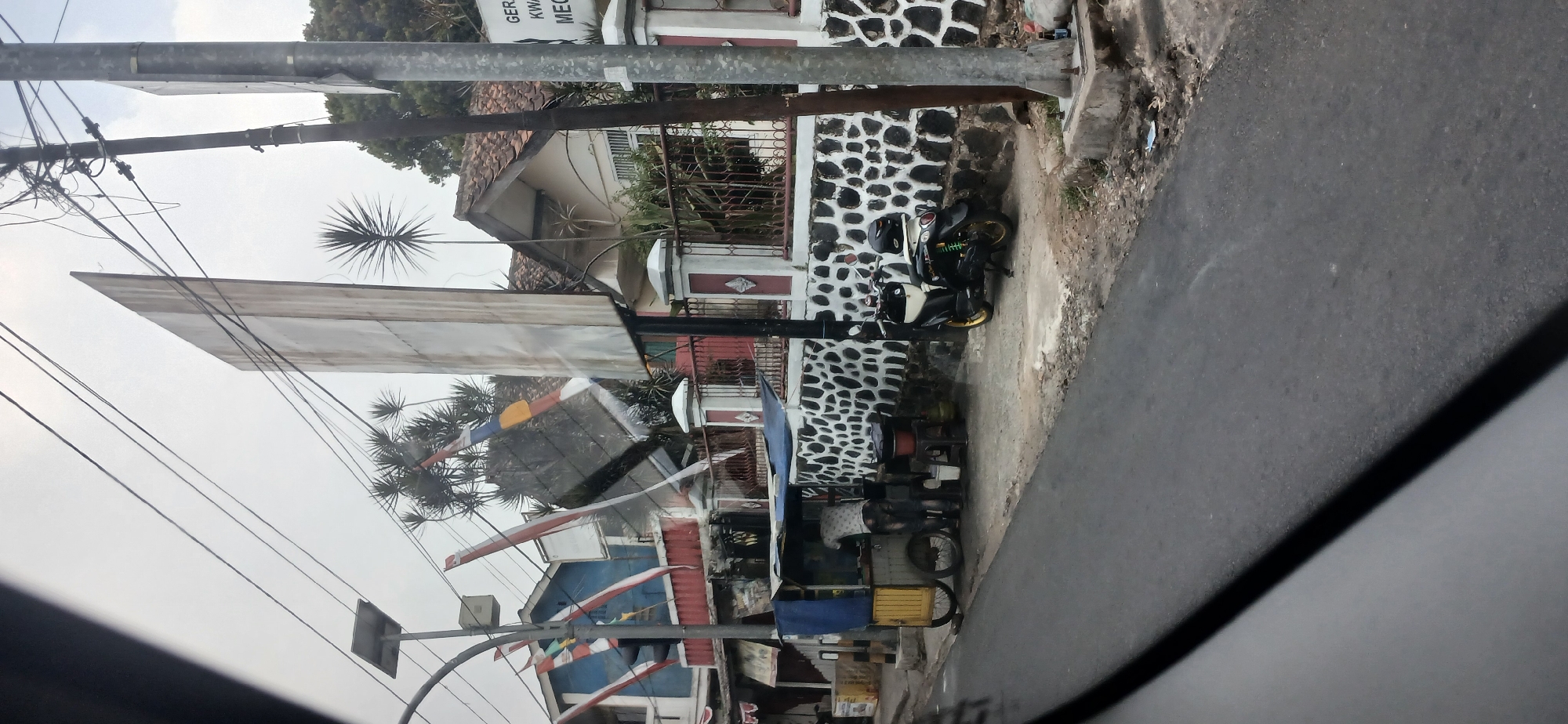 The image depicts a street scene in Megamendung, Indonesia, characterized by a cloudy sky and a mixture of residential and commercial structures. In the foreground, an asphalt road is visible, with a dark shadow cast across it. To the right, a utilitarian metal utility pole stands tall, supporting a dense network of electrical wires. Adjacent to the pole, a white and red striped fence encloses a property with a house featuring a tiled roof and barred windows.
The focal point of the scene is a food cart situated on the sidewalk. The cart is yellow with a blue awning and is manned by a person wearing a light-colored shirt with a patterned design, who appears to be attending to customers or preparing food. A gas tank is visible near the cart. A black scooter with yellow accents is parked beside the cart, with a black helmet resting on its seat.
Further back, a building with blue and red accents is partially visible, adorned with Indonesian flags. A large, irregularly shaped billboard stands prominently in the background, angled precariously. Trees with spiky foliage are scattered throughout the scene, some of which appear to be palm trees. The overall impression is one of a typical street in a developing urban or semi-urban area.