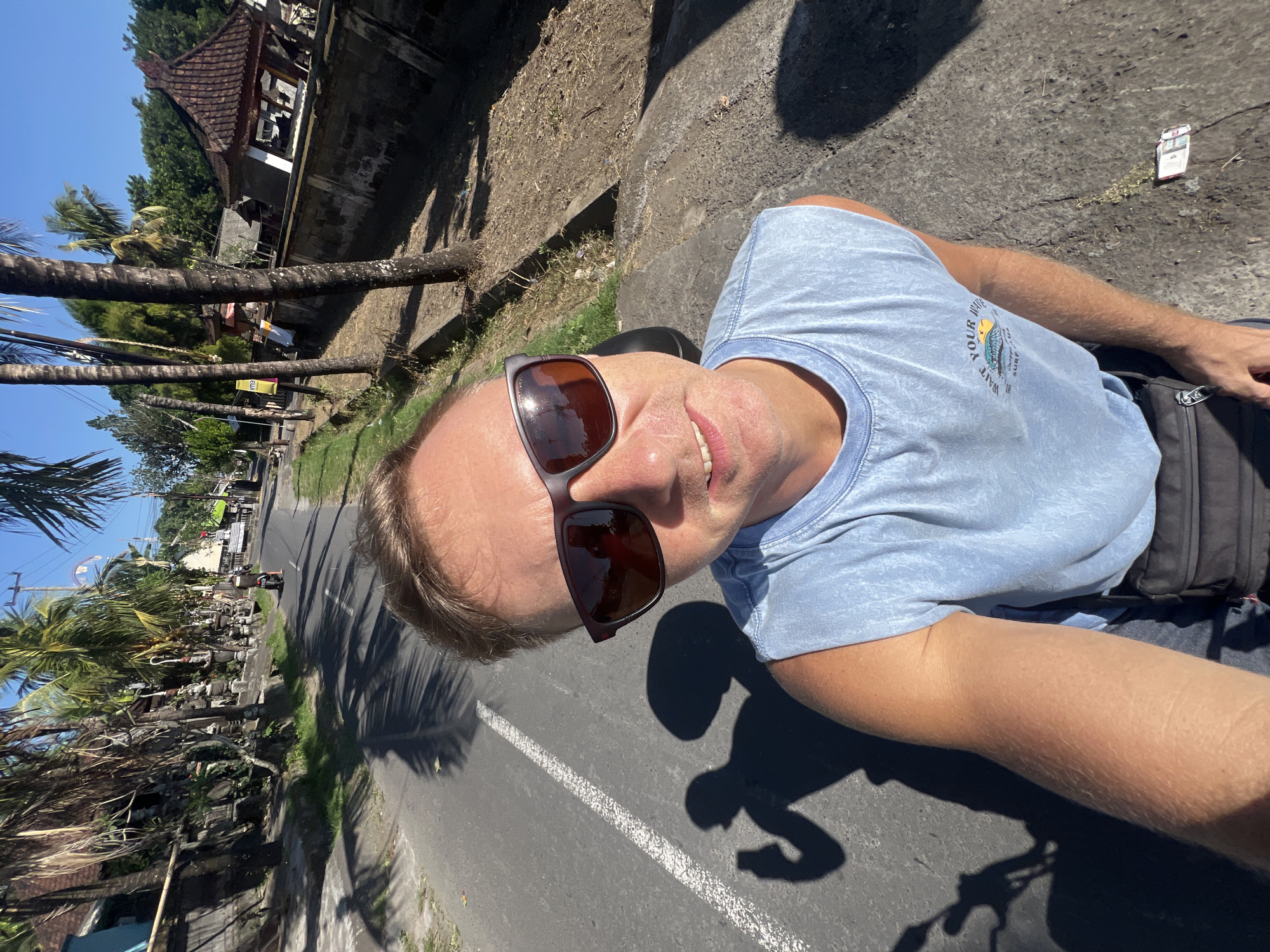 A man wearing a light blue sleeveless t-shirt and sunglasses takes a selfie on a street in Sukawati, Indonesia. The man is smiling and his right arm is extended, holding a phone that is not visible. He is wearing a dark fanny pack around his waist. The background shows a road with a white dashed line, palm trees, and buildings with traditional Indonesian architecture. A shadow of the man on a scooter is visible on the road to his left. The sky is clear and blue.