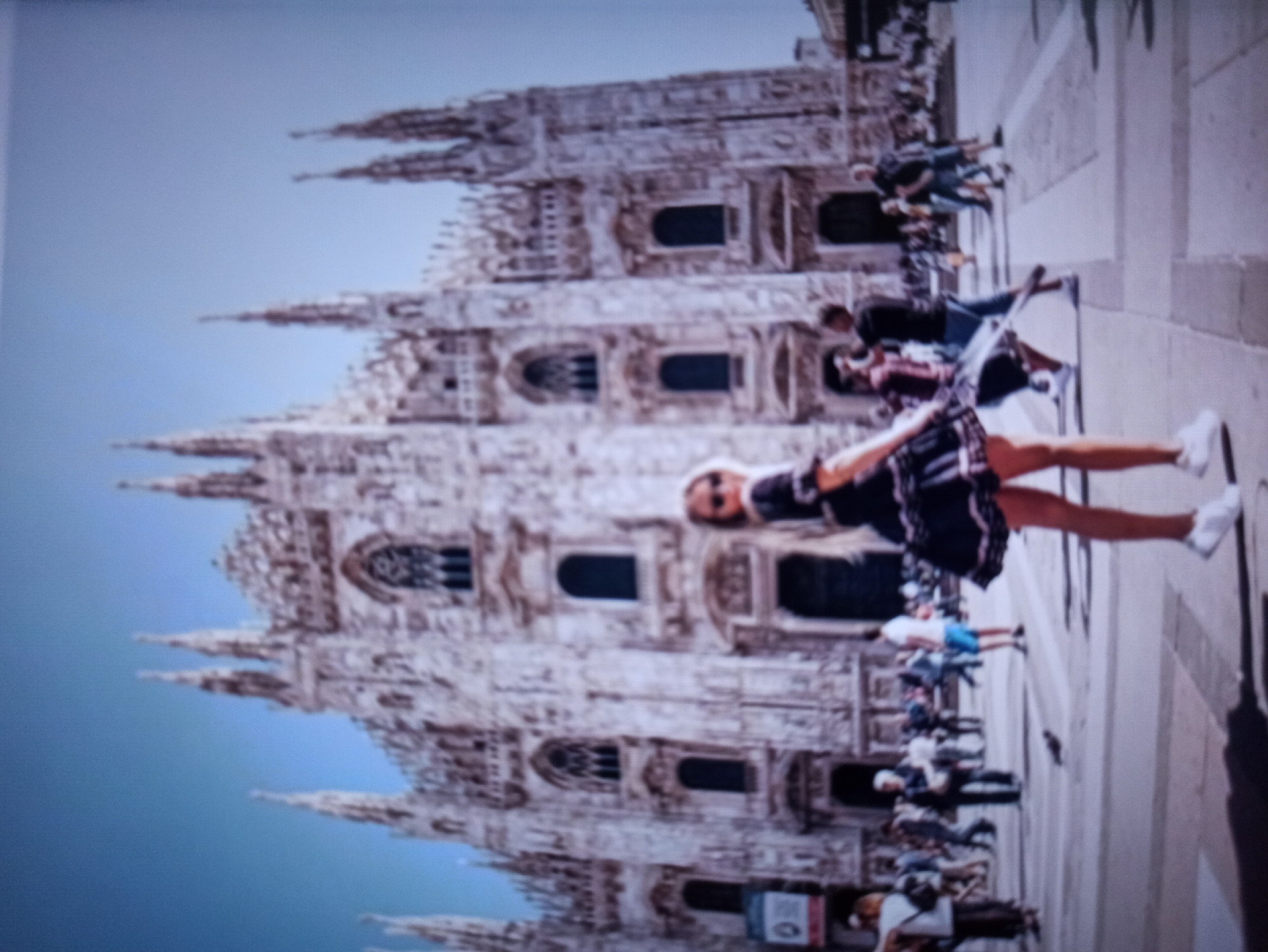 The image depicts a wide-angle shot of the Milan Cathedral in Italy, with a young woman in the foreground. The woman is wearing a black dress with a tiered skirt and white sneakers, and her blonde hair is visible. She is positioned on a paved square in front of the cathedral. Several other people are scattered across the square, some walking, some standing. The cathedral itself is a large, ornate Gothic structure with numerous spires and intricate details. The sky above is a clear blue. The image is slightly blurry, which gives it a soft, impressionistic quality. The city of Balakliya, Ukraine is not depicted in this image.
