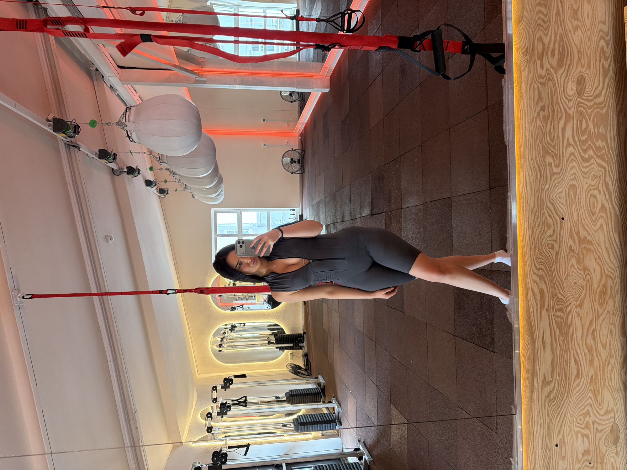 A woman wearing a gray bodysuit and white socks is posing for a selfie in a gym. She is holding a cell phone in her right hand and looking into the camera. The gym appears to be in Moscow, Russia, based on its architectural style and design elements. The gym features modern exercise equipment, including weight machines, suspension training systems, and large white exercise balls suspended from the ceiling. The floor is dark brown and tiled. The walls are painted white, and there are mirrors on both sides of the gym, reflecting the exercise equipment and the woman. There is a wooden panel with a light strip running along its edge on the right side of the frame. Windows are visible in the background, providing natural light. The overall atmosphere of the gym is bright and modern.