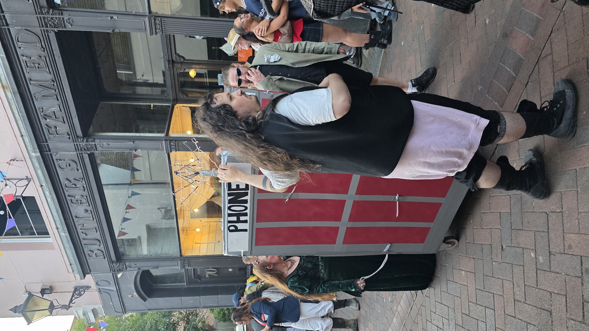 In St Leonards, United Kingdom, an outdoor scene unfolds on a brick-paved street in front of a building labeled "BUTLERS FAMED." A man with long, curly brown hair, wearing a black vest over a white t-shirt and a pale pink skirt, is pushing a mobile red and silver structure resembling a phone booth. The structure has "PHONE" written in black letters on a white sign at the top. To the left of the phone booth, a woman in a long, dark green velvet dress and a necklace stands, holding a white rope attached to the phone booth. Other individuals are visible in the background, some appearing to be dressed in costumes. The building has a dark facade with large windows and an ornate entrance. Bunting is strung across the building's facade.