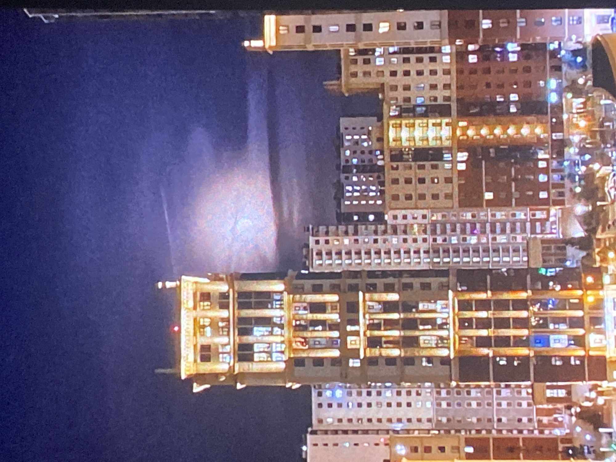 The image shows a portion of the cityscape of Saint Petersburg, Russia, at night. Prominent buildings are visible, with numerous illuminated windows suggesting residential or commercial occupancy. A significant light source in the sky, possibly the moon or another strong light source, casts a glow across the scene. The buildings exhibit various architectural styles and heights.