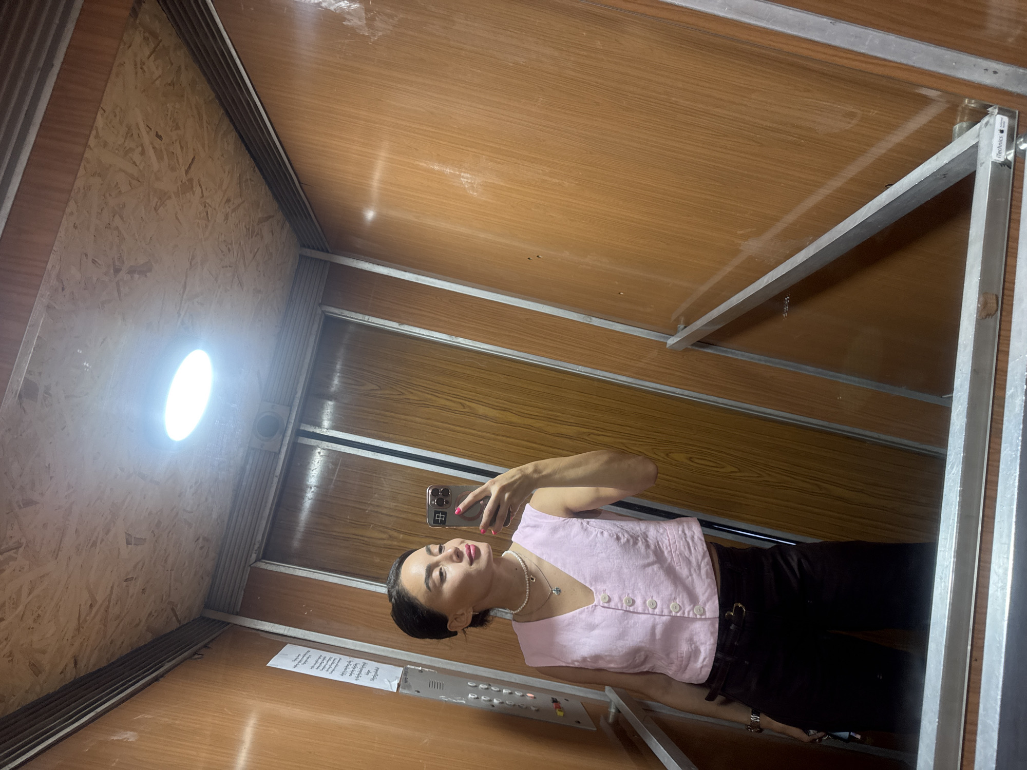 An Asian woman is taking a mirror selfie in an elevator. She is wearing a pink vest and dark pants. The elevator walls are made of wood paneling, and the ceiling is covered in particle board. There is a light fixture in the ceiling. A paper notice is affixed to the elevator wall. The elevator is located in the city of Tbilisi, Georgia.