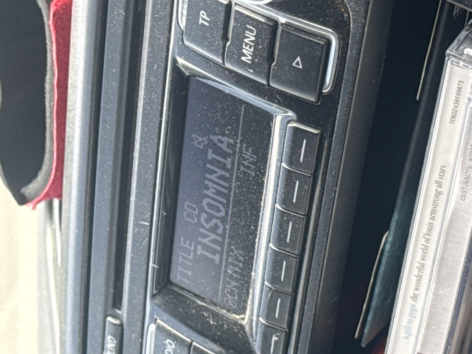 The image presents a detailed, close-up view of the interior of a vehicle, specifically focusing on a car's dashboard-mounted radio unit and a storage compartment beneath it.
The central feature is an illuminated digital display on the car radio, which reads "TITLE CD" on the upper line and "INSOMNIA" prominently in larger characters on the second line. A third line of text below "INSOMNIA" is partially legible, beginning with "SCN MIX" and "INF". To the left of the display, partial button labels "OUND" and "DIO" are visible. To the right, a vertical stack of control buttons is present, labeled "TP", "MENU", and a triangular symbol pointing right. A horizontal row of six identical rectangular buttons, each marked with a horizontal dash, is situated directly beneath the display. The entire radio unit, including the display bezel and physical buttons, is covered in a noticeable layer of fine dust and small particulate matter.
Below the radio unit, a storage slot contains a compact disc (CD) case. Text visible on the spine or edge of this case includes "a gift to pops the wonderful world of louis armstrong all stars". Further along the case, a numerical sequence "00602438599875" is evident, along with an alphanumeric code "CDSTUMM275, 50000...".
In the upper right portion of the image, resting on the dashboard surface above the radio, a red object, possibly a pair of sunglasses with red frames or a piece of red fabric, is partially visible. The dashboard surface surrounding the radio also shows accumulated dust.
The specific location in the city of Eisenerz, Austria, is not verifiable from the visual information provided in the image.