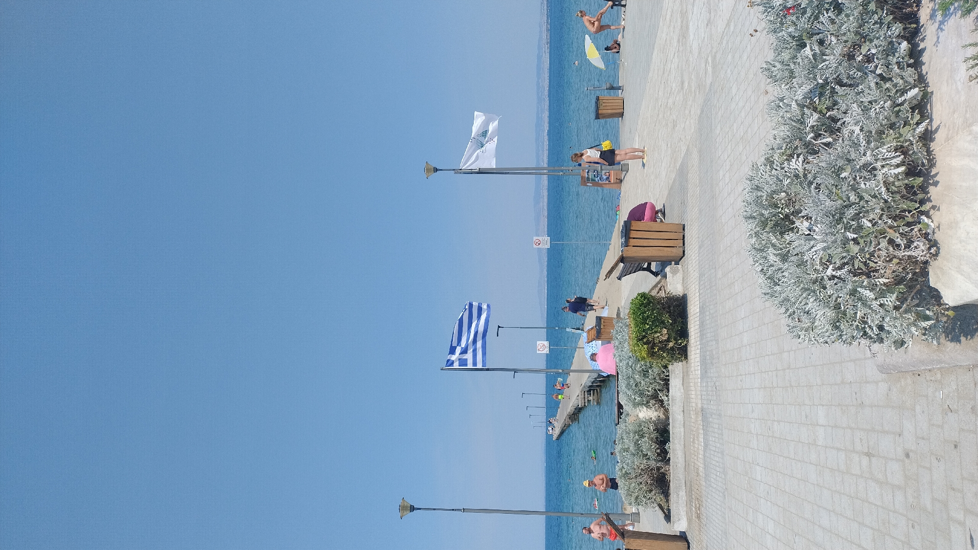 In Perea, Greece, a wooden pier extends into the calm blue sea under a clear sky. A large Greek flag waves prominently from a flagpole on the pier. Another white flag with a logo flies nearby. Several people are visible on the pier, some walking and others seated. A few individuals are in the water, and others are on the paved walkway beside the sea, where there are benches and planters with greenery. The walkway is made of light-colored paving stones. The background shows distant hills or land.