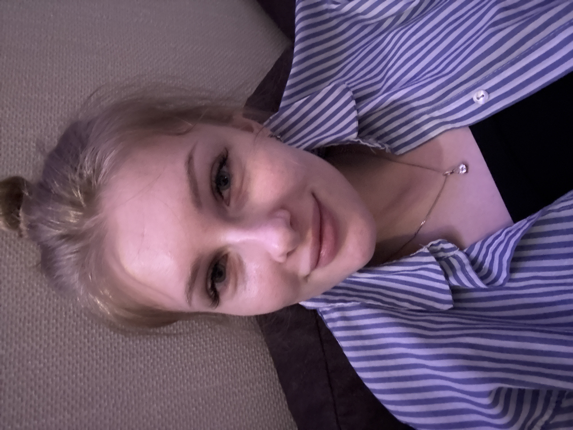 A young woman with blonde hair pulled back in a bun is lying down, possibly on a couch, wearing a blue and white striped button-down shirt over a black top. She has light skin, green eyes, and appears to be smiling slightly. A delicate necklace with a pendant is visible around her neck. The background includes a beige textured surface. The location, Perm, Russia, cannot be determined from the image.
