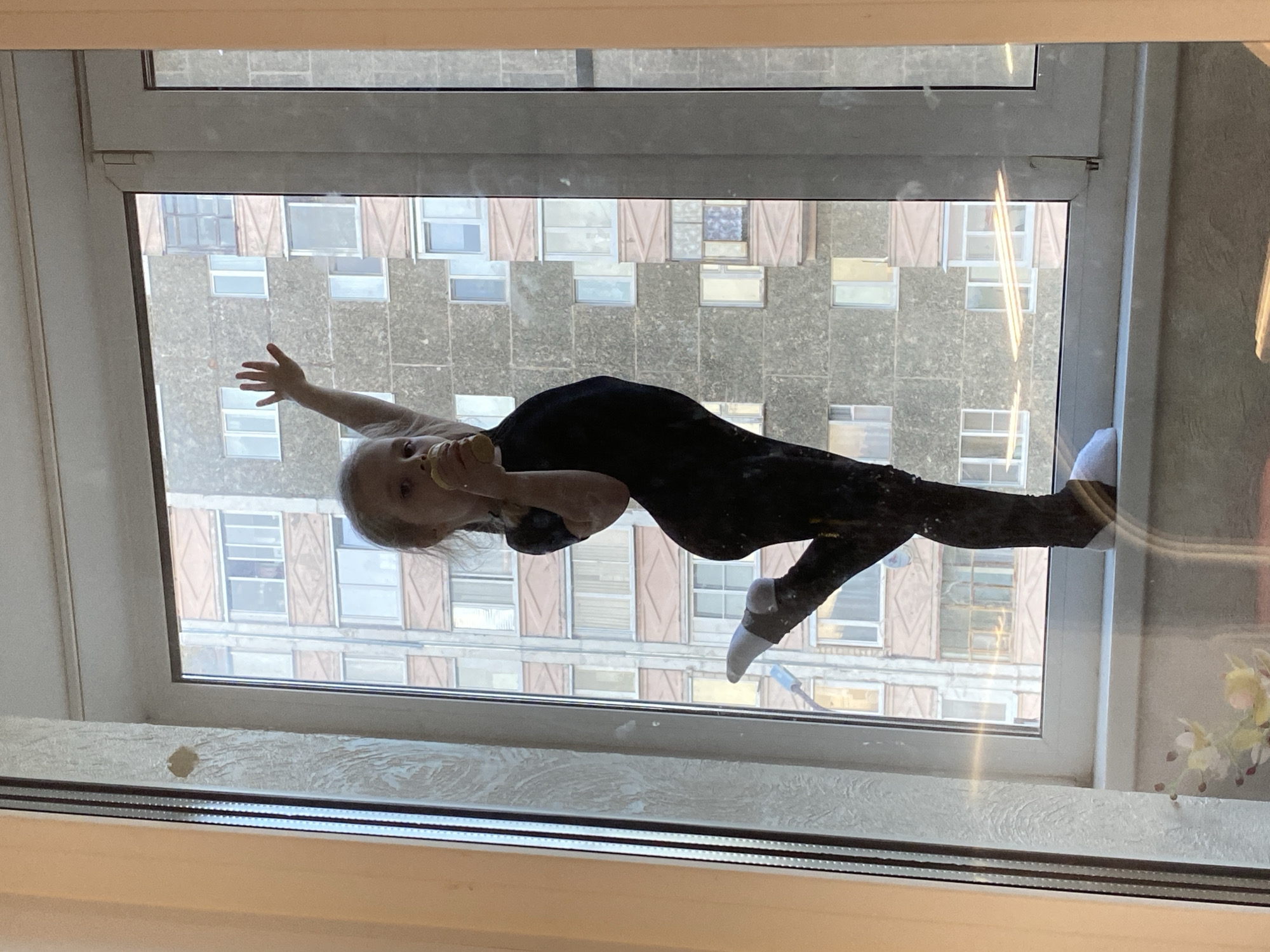 A child is visible through a window. The child is wearing a black leotard and white socks, and is reaching out with one hand while holding something in the other. Outside the window, a large, grey building with multiple windows, some with brown shutters, is visible. Given the information about the location being Votkinsk, Russia, it's likely that the architecture and building style are representative of that area. The window frame is white and the room behind the window is softly lit.