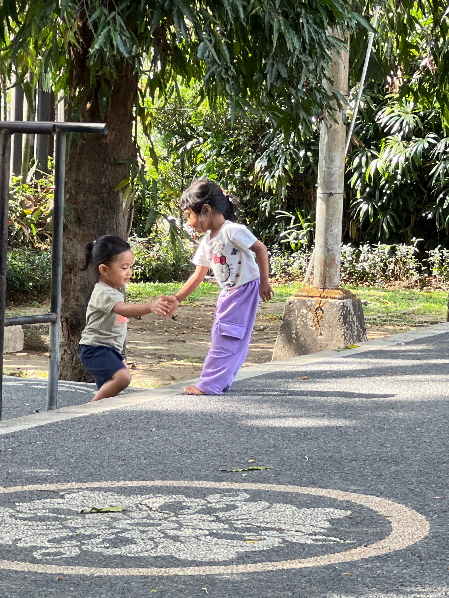 The image depicts two young girls, one older and one younger, interacting outdoors in a park-like setting, possibly in Denpasar, Indonesia, given the presence of lush tropical vegetation. The older girl, wearing a white t-shirt with a Hello Kitty graphic and purple pants, is extending her hand to the younger girl. The younger girl, dressed in a tan t-shirt and dark shorts, is reaching out to hold her hand. They appear to be standing on a paved surface near a concrete structure and a tree. The background is filled with dense green foliage, suggesting a sunny day in a tropical environment. The ground in the foreground shows a textured asphalt surface with white markings, possibly part of a playground or recreational area.