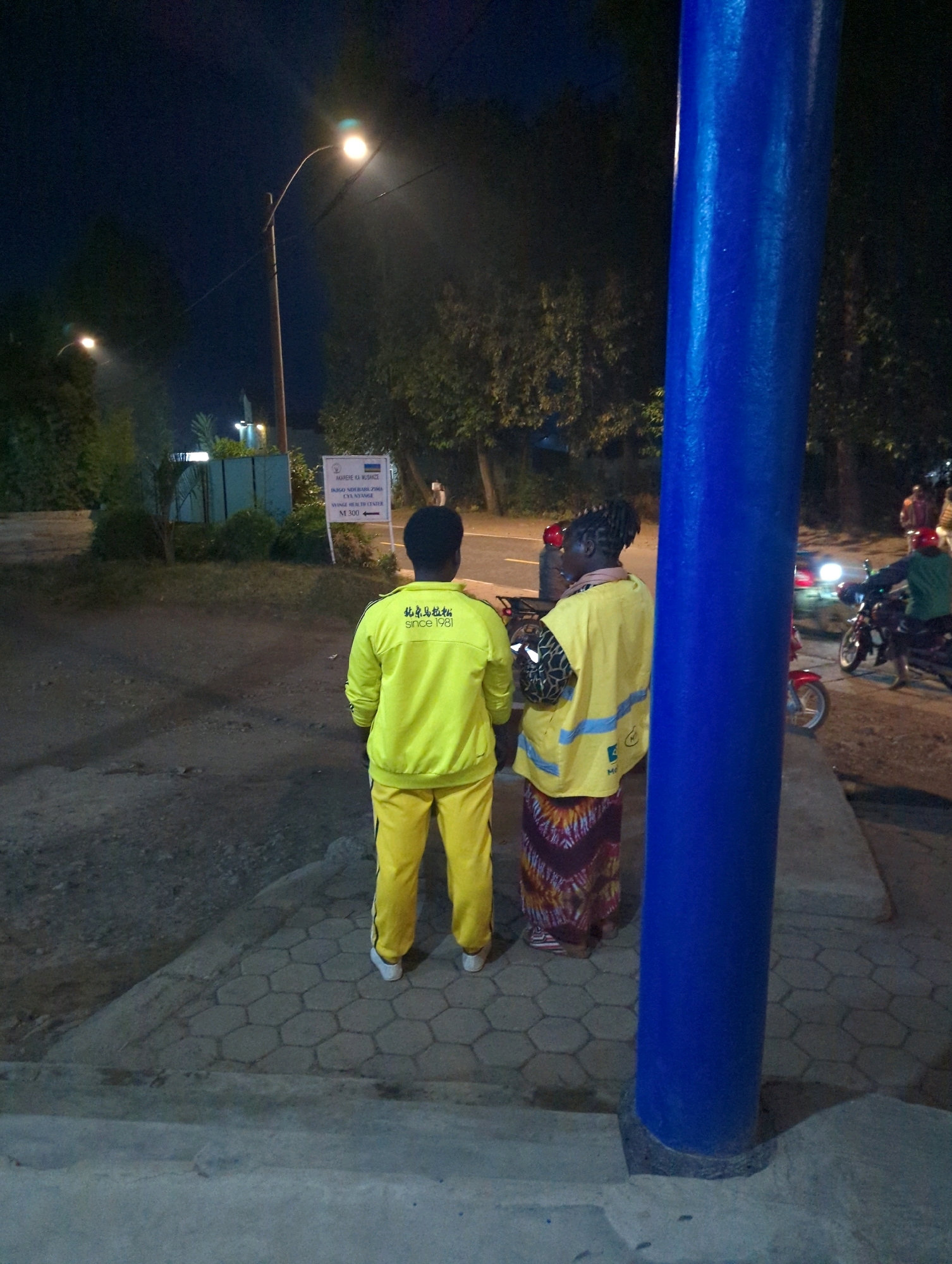 At night in Musanze District, Rwanda, two individuals stand on a sidewalk next to a large blue cylindrical pillar. The person on the left, viewed from the back, wears a bright yellow tracksuit with a logo on the back of the jacket featuring Chinese characters and the text "Since 1981." The person on the right, also viewed from the back, wears a yellow vest over a patterned skirt. In the background, a street light illuminates a grassy area with trees and a signpost. Motorbikes are visible on the road, with one rider wearing a green jacket and a red helmet. The scene appears to be near a health center, as indicated by the sign. The ground is paved with hexagonal concrete blocks and asphalt.