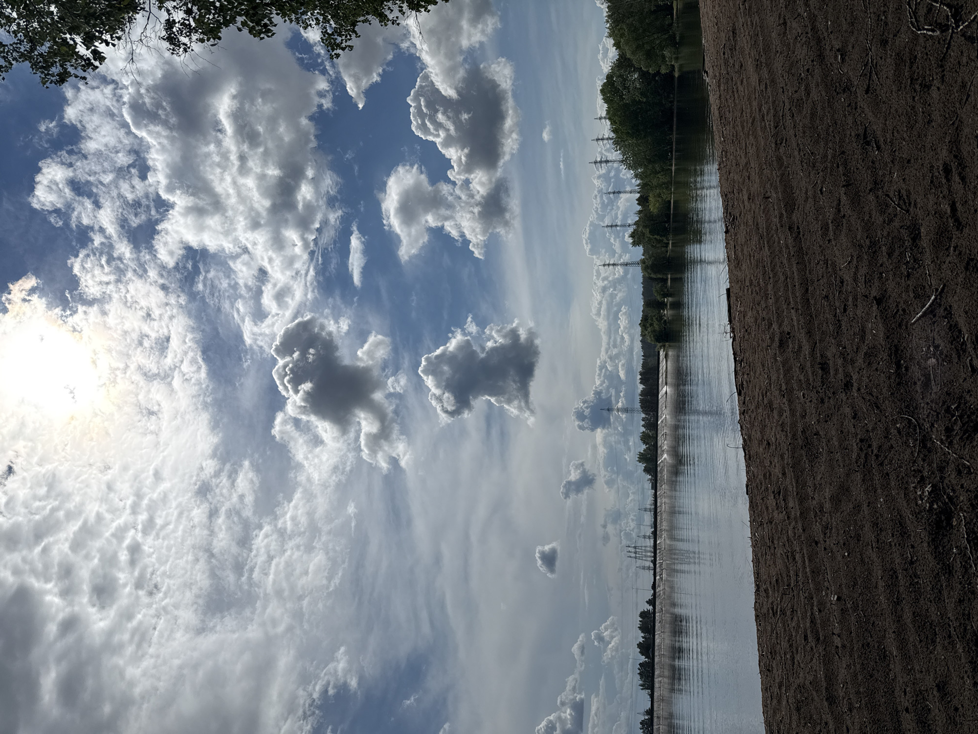 The image presents a landscape scene with a body of water, likely a river or lake, flanked by foliage and land. A strip of dark soil or dirt occupies the right foreground, while the water surface reflects the sky and surrounding vegetation. Overhead power lines or pylons can be seen in the distance alongside trees. Cumulus clouds dominate the sky with patches of blue in between. The sun is visible on the left side. The photograph is taken at an angle. It depicts a natural environment with signs of infrastructure. Given the mention of "Noviy, Russia", it is likely this is a landscape found in or around that city.