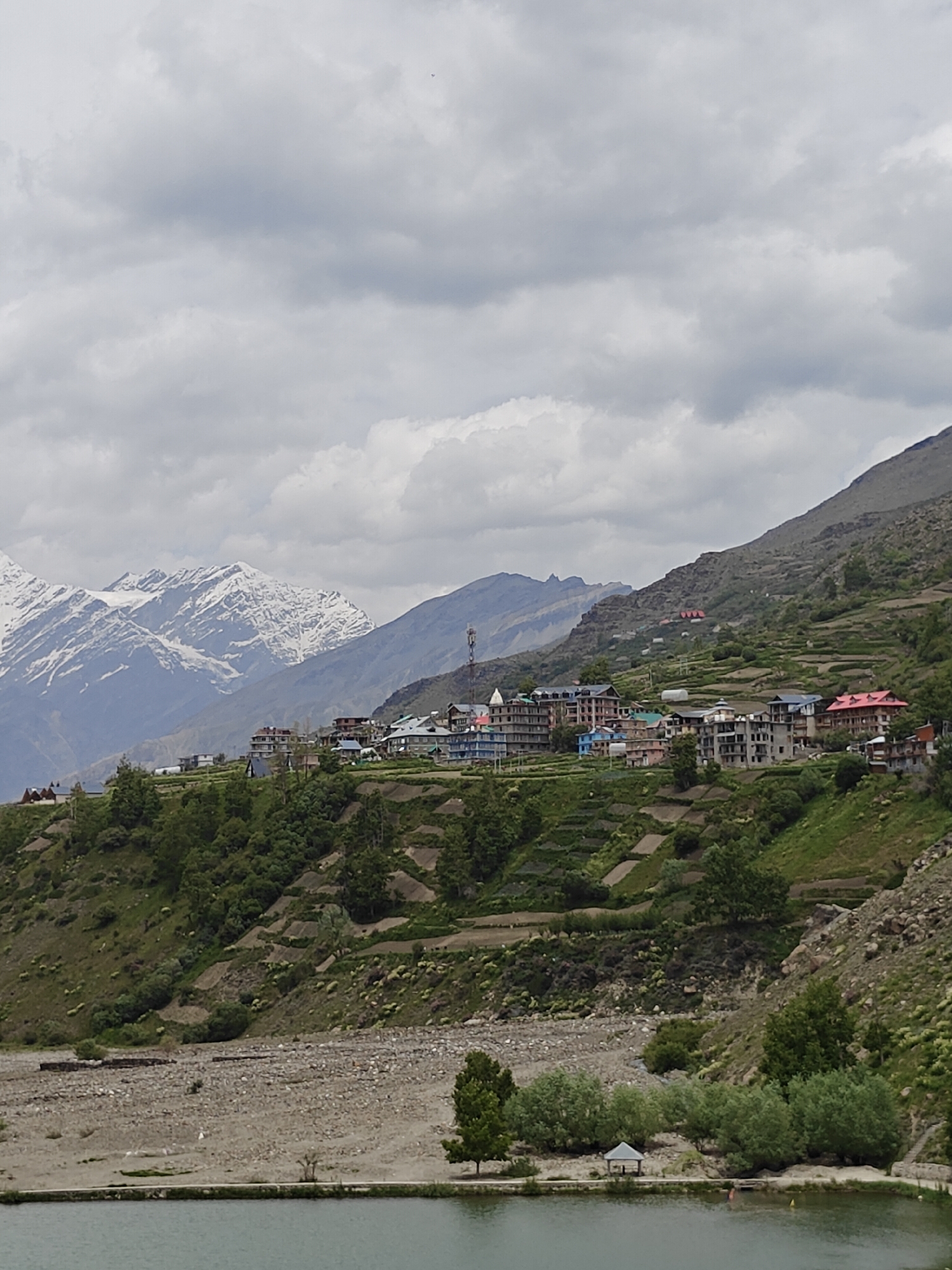 This image depicts a panoramic view of Yamling, India, a scenic town nestled amidst the Himalayan mountains. The foreground features a tranquil body of water, likely a lake, reflecting the overcast sky. Beyond the water, a rocky terrain gives way to a verdant hillside dotted with terraced fields and lush green trees. A cluster of houses with colorful roofs, characteristic of the region's architecture, are perched on the slopes, forming a quaint village. In the background, majestic snow-capped mountains rise, their peaks shrouded in clouds, adding to the breathtaking natural beauty of the landscape. The overall scene is one of serene rural life, harmoniously coexisting with the imposing grandeur of the Himalayas.