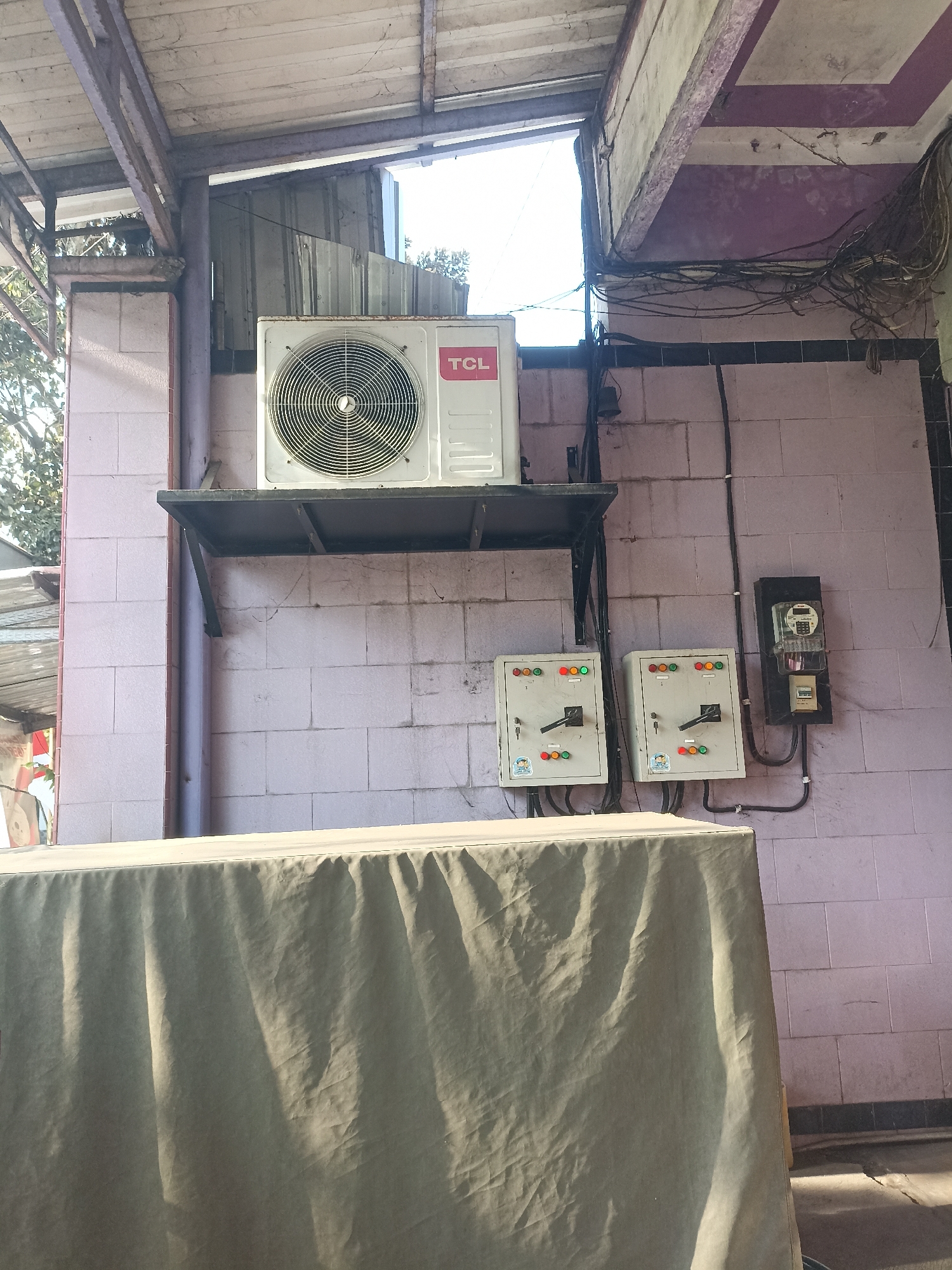 The image shows an outdoor area in Tompokersari, Indonesia, with an air conditioning unit mounted on a metal shelf against a light pink tiled wall. To the right of the air conditioning unit are two grey electrical breaker boxes with visible switches and indicator lights. Further to the right, a black electrical meter is mounted on the wall. Above the electrical boxes and air conditioner, numerous black electrical wires are bundled together. In the foreground, a large object covered with a light grey cloth dominates the lower half of the image. The scene appears to be a service area or an outdoor portion of a building.