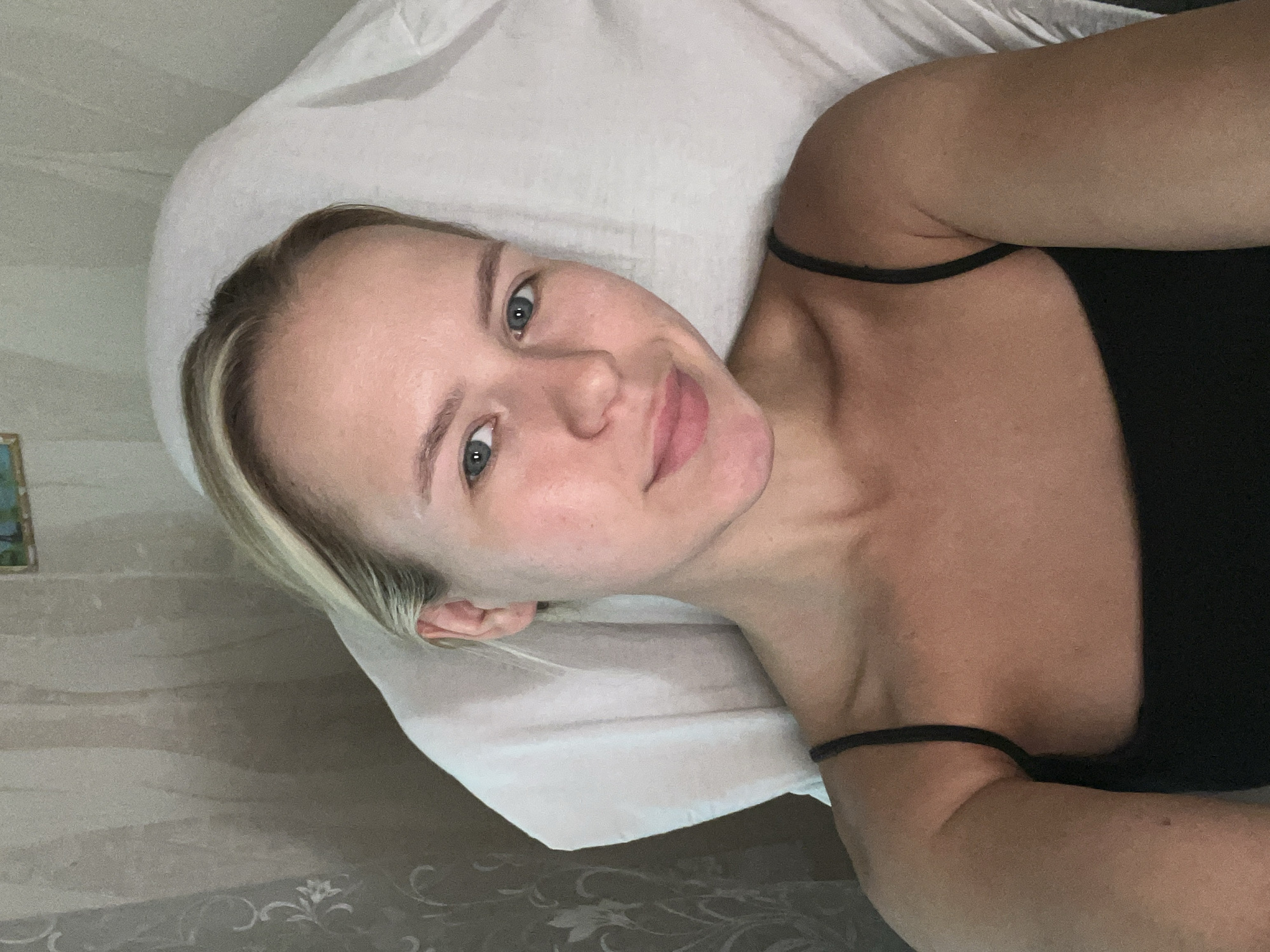 A fair-skinned woman with blonde hair and blue eyes is pictured in a close-up selfie. She is lying down, resting her head on a white surface that appears to be a pillow or a headrest. Her hair is pulled back, revealing her face and ears. She is wearing a black camisole. The background shows a light-colored curtain with a subtle floral pattern. A small framed picture is visible on the wall behind her, partially obscured by the curtain. The overall lighting suggests an indoor setting.