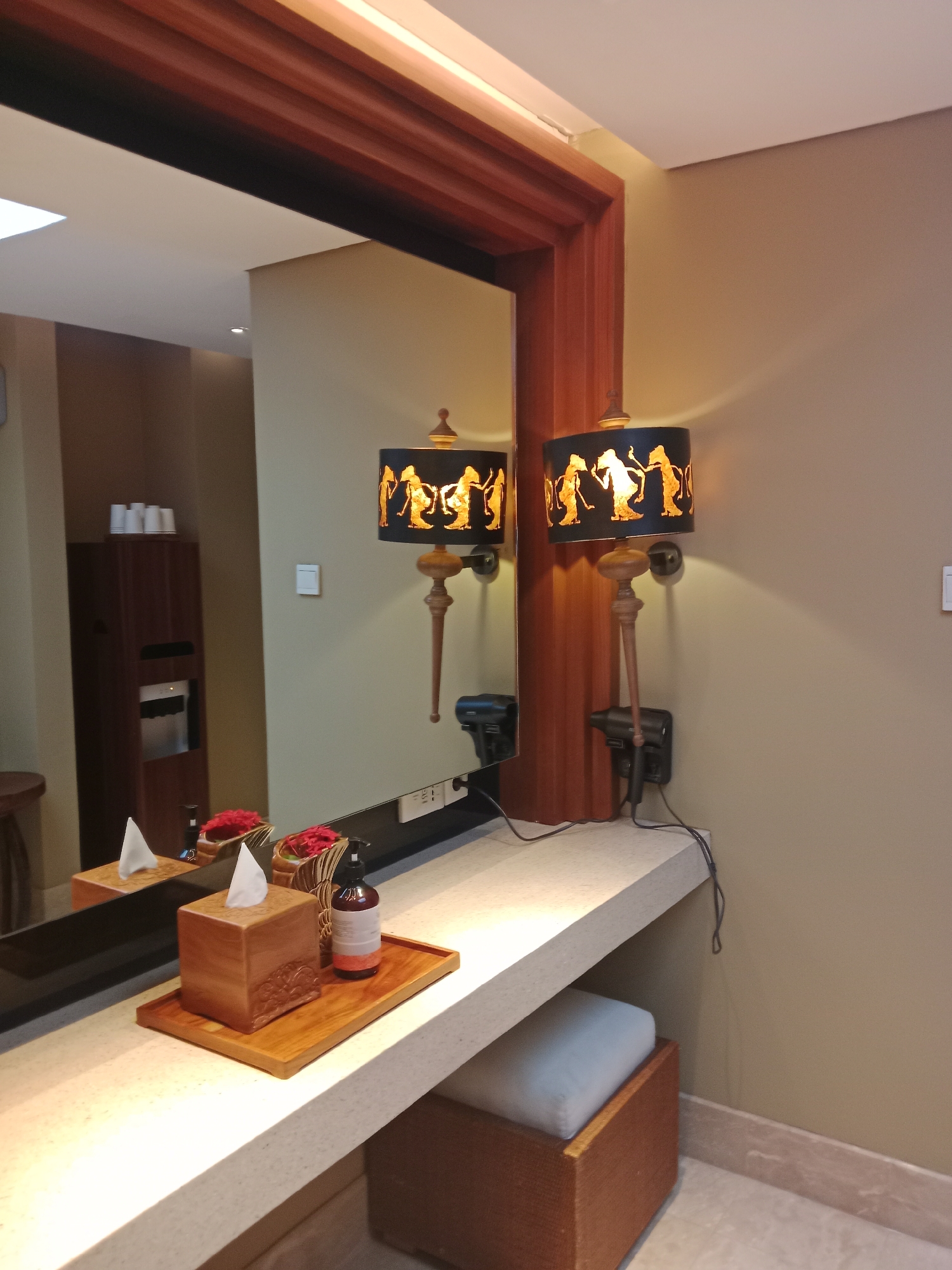 This image depicts a vanity area, likely within a hotel room or spa, situated in Kuta Selatan, Indonesia. The scene is dominated by a large mirror reflecting a wooden-framed area above and a wall-mounted lamp with a decorative shade featuring golden silhouettes of figures. In front of the mirror is a countertop displaying a wooden tissue box, a small wooden tray holding red decorative flowers, and a dark bottle with a dispenser cap. A hairdryer is mounted on the wall below one of the lamps. To the left, a tall wooden cabinet is visible, with a water dispenser and stacked cups on top. A small, cushioned stool with a woven base is positioned beneath the countertop on the right. The walls are painted a neutral, light tan color, and the floor appears to be tiled. The lighting suggests an indoor setting, possibly during the day or with artificial illumination.