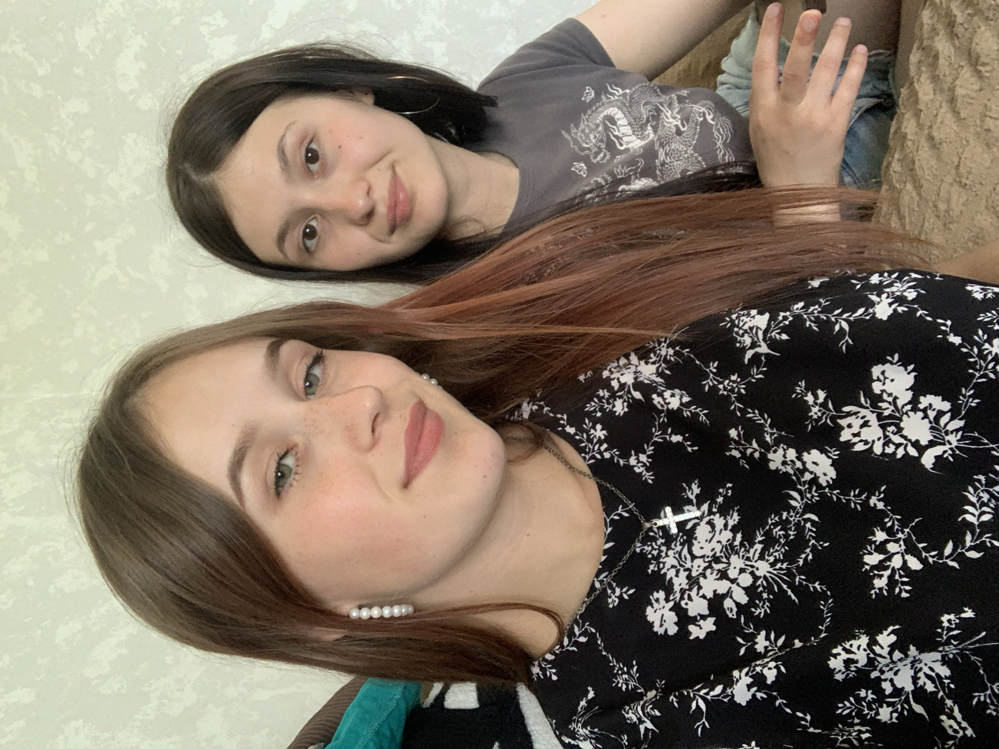 Two young women are posing closely together for a photo indoors. The woman in the foreground is wearing a black dress with a white floral pattern and a silver cross necklace. She has brown hair with lighter reddish-brown highlights and is wearing small pearl earrings. The woman behind her is wearing a gray t-shirt with a graphic print and is lying on a brown textured surface. She has dark brown hair. The background is a light-colored, textured wall. The city of Фоки, Russia is not discernible in this image.