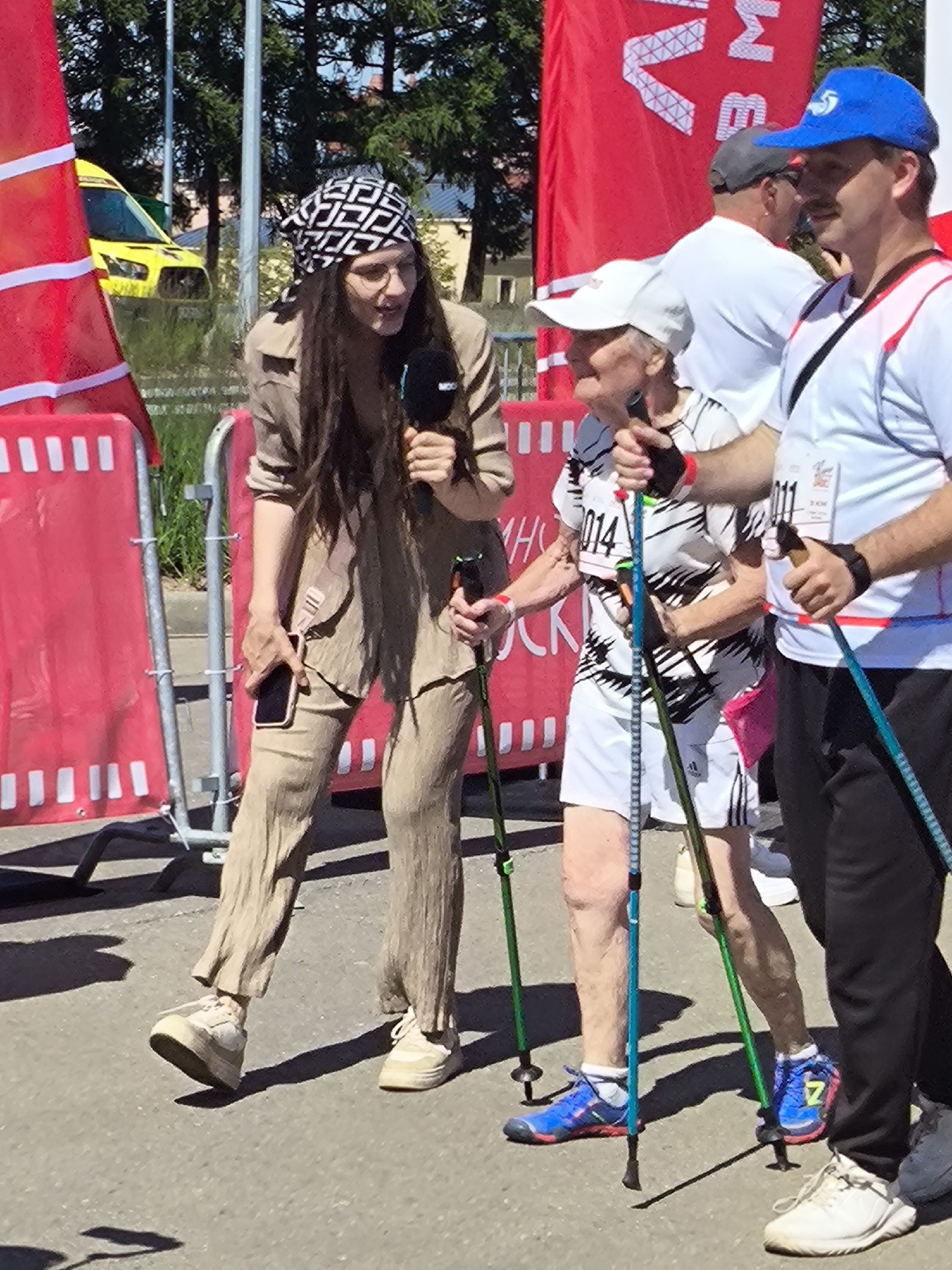 In Moscow, Russia, a woman with long dark hair, wearing glasses and a bandana, is holding a microphone and speaking. She is dressed in a tan colored outfit consisting of a jacket and loose-fitting pants. Next to her, an elderly person in a white and black striped shirt and white shorts is holding Nordic walking poles. To their right, a man in a white t-shirt and blue baseball cap is also holding a Nordic walking pole. Behind them, other people are visible, and a yellow ambulance is parked in the background. The event appears to be outdoors, possibly a race or athletic competition, with red banners and barriers set up.