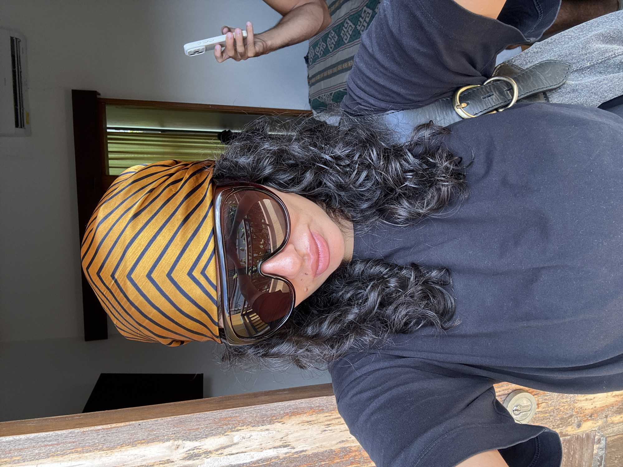 The image shows a person wearing a black top and a gold and black patterned headscarf, with large sunglasses, standing in what appears to be a building with wooden railings. In the background, there is a person holding a white smartphone, standing next to a patterned cushion. The location is inferred to be in North Kuta, Indonesia.