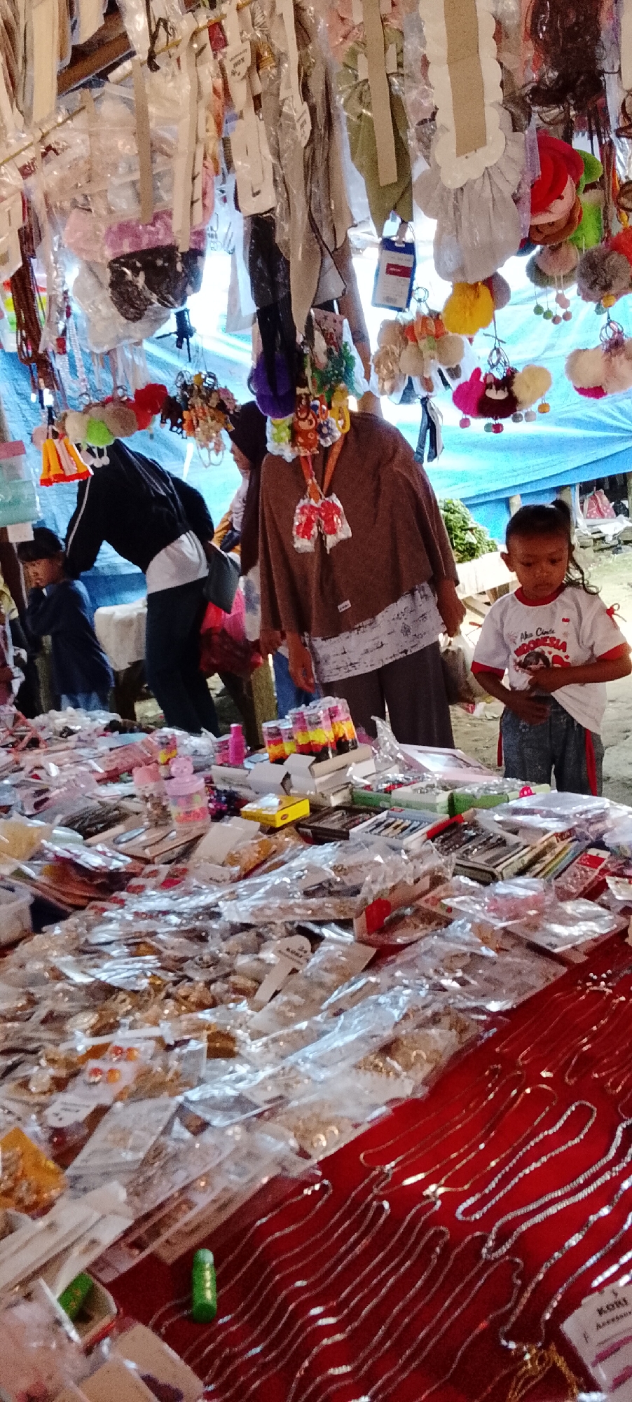 The image depicts an outdoor market stall, likely in Bahway, Indonesia, filled with an assortment of goods. In the foreground, a table is laden with various items, many packaged in clear plastic, including what appear to be jewelry, hair accessories, and small trinkets. A child in a white t-shirt with red trim and gray patterned shorts stands to the right, looking down at something in their hands. Behind them, a person in a brown top and patterned skirt is visible, also looking towards the table. Another person, wearing a dark jacket over a white shirt and dark pants, is partially obscured on the left, bending down. The stall is covered by a blue tarp, and various items, including keychains and decorative objects, hang from the overhead structure. The overall impression is one of a bustling, informal marketplace.