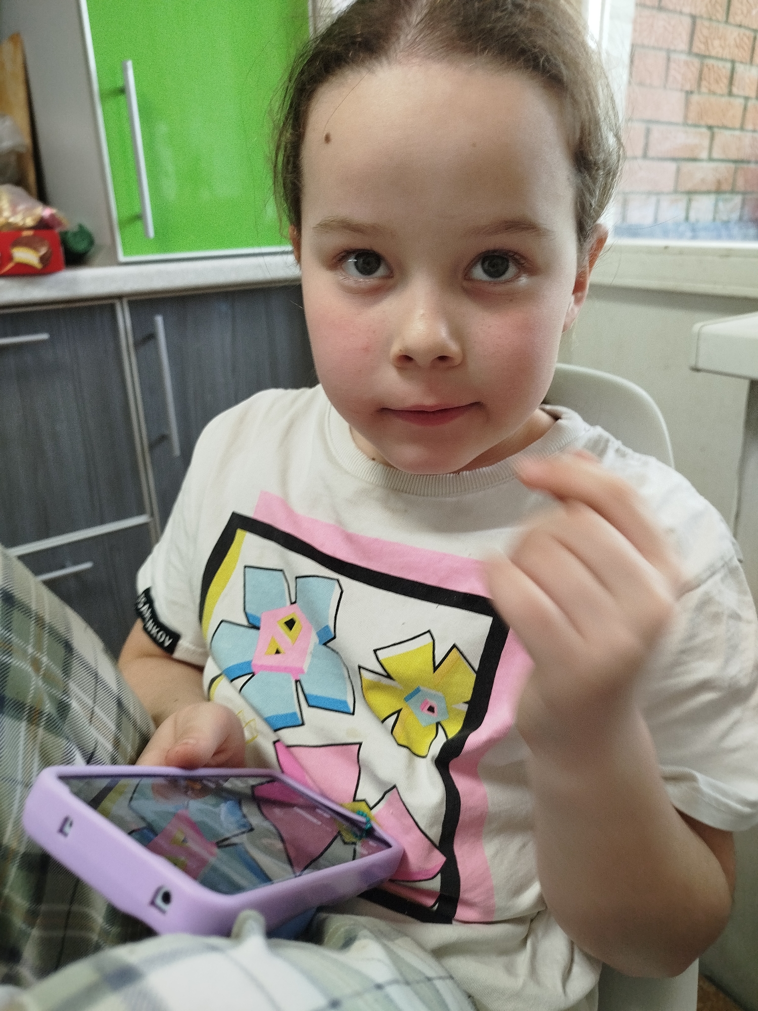 A young girl with fair skin and light brown hair pulled back is sitting in a chair, holding a lavender-cased mobile phone. She is wearing a white t-shirt with a graphic design featuring stylized flowers outlined in black and pink, yellow, and blue colors.
The girl is indoors, possibly at a kitchen or dining area. Behind her is a green kitchen cabinet with a silver handle and a set of gray cabinets. A brick wall is visible through a window in the background. The location is Chaikovsky, Russia.