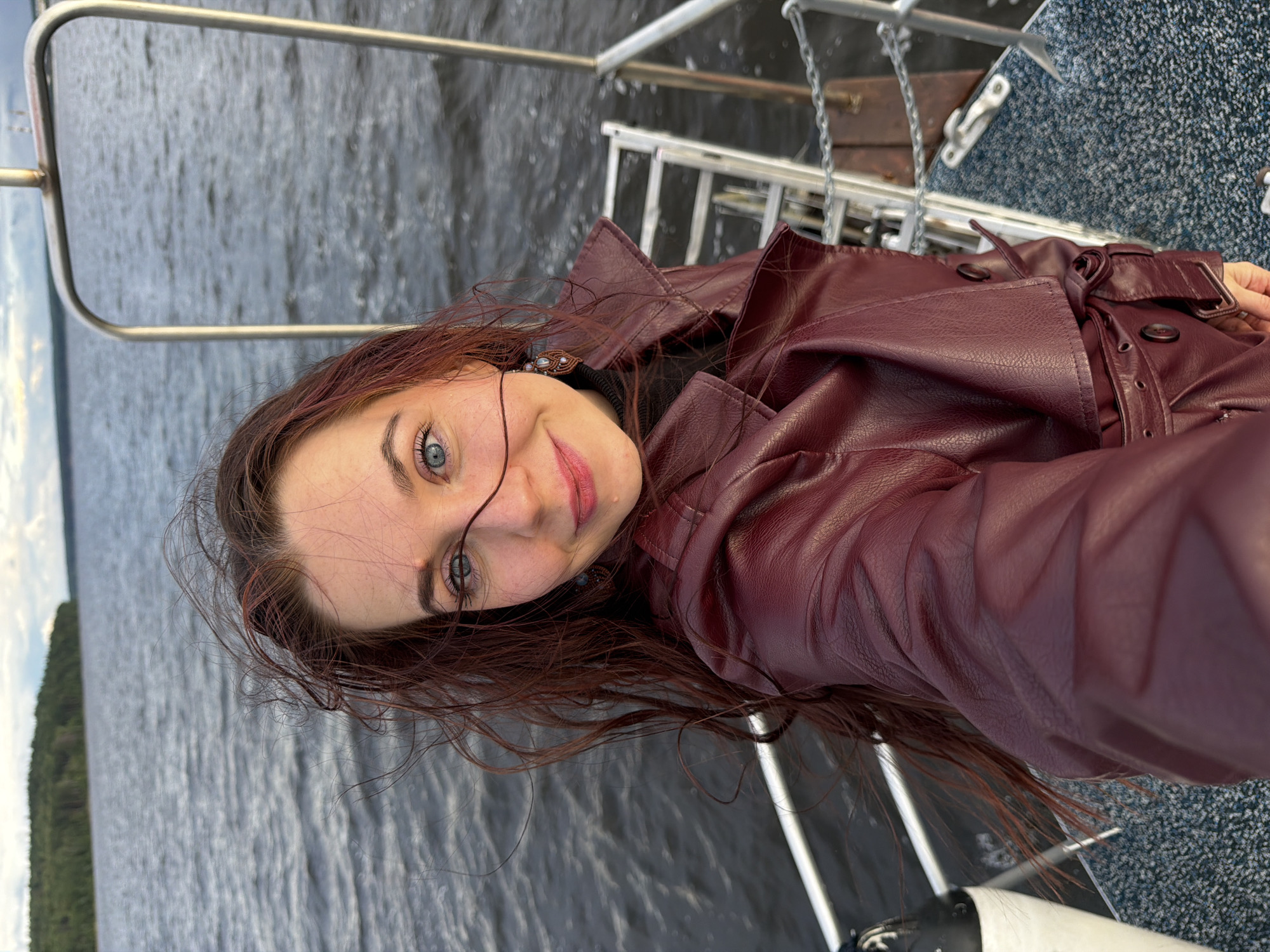 A young woman with long, auburn hair stands on the deck of a boat, taking a selfie. She is wearing a dark red leather trench coat over a black turtleneck. The boat is on a body of water, possibly the Kama River, near the city of Chaikovsky, Russia. The water is dark and choppy, and the sky is overcast with some clouds. In the background, a tree-lined shore is visible. The woman has bright blue eyes and a faint smile. Her hair is windswept, with strands blowing across her face. The boat has metal railings and a textured deck.