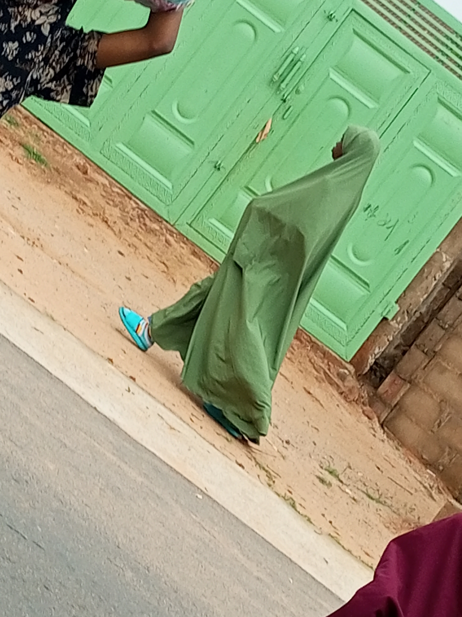 A person, partially visible from the shoulder up and wearing a black and white patterned garment, stands to the left, holding a pink and blue object. To the right, another person, facing away from the camera and covered in a flowing olive green garment and headscarf, walks across a dirt path. This individual is wearing a bright turquoise sandal on their left foot. Behind them, a large, textured, light green gate with visible paneling and decorative molding dominates the background. The gate appears to be made of wood. The ground is a mix of dry, light brown dirt with some scattered green vegetation and pebbles. To the right of the dirt path, a section of a brick wall is visible. The overall scene suggests an outdoor setting, possibly a residential area, given the presence of a gate and wall. The image is taken from a slightly elevated, angled perspective, looking down and towards the right. The context of Tempure, Nigeria, is not evident from the visual information in the image itself.