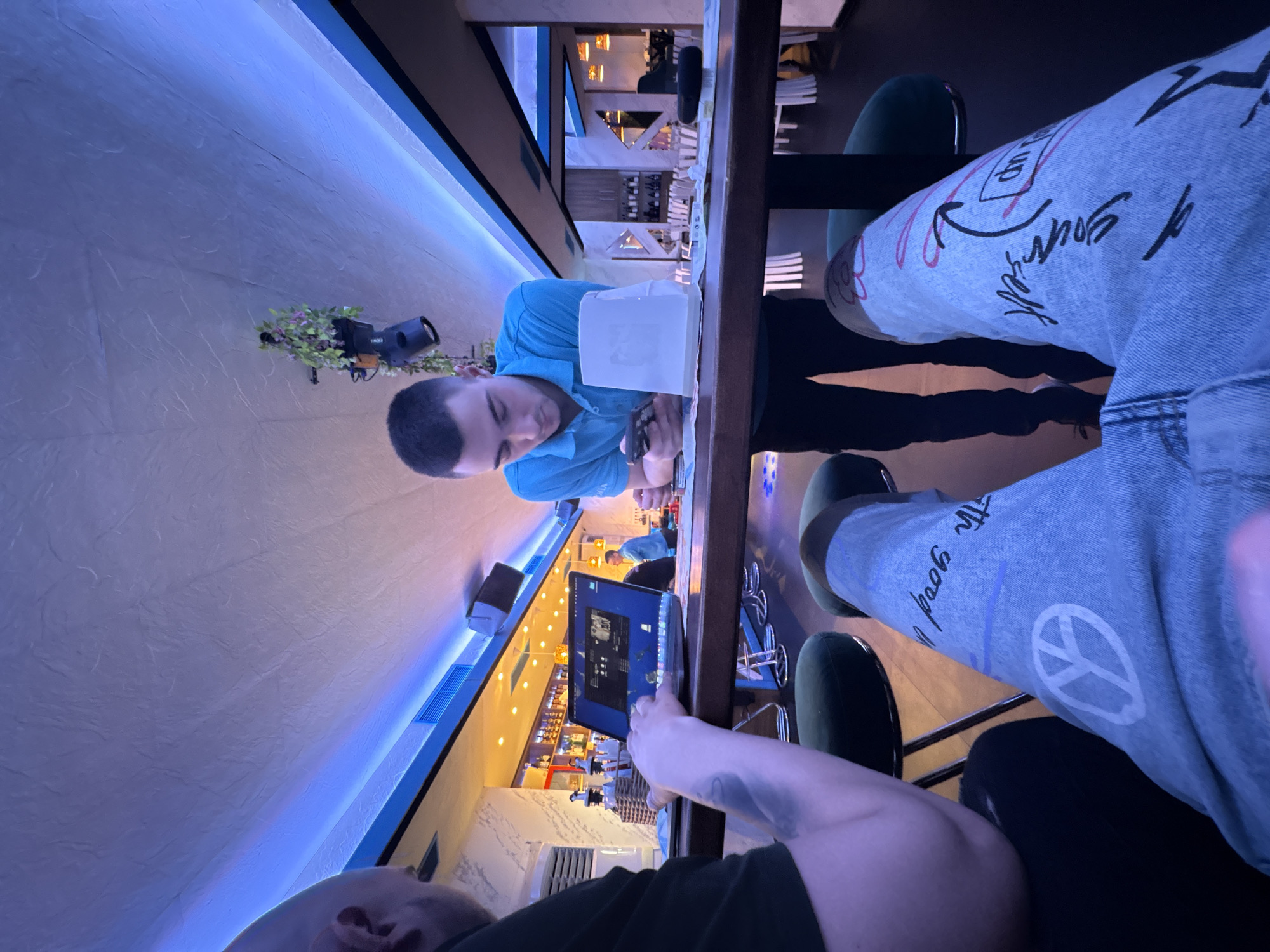 The image depicts an indoor scene in what appears to be a modern restaurant or bar, characterized by dim, ambient lighting, likely during the evening or night. A prominent feature is a continuous strip of blue LED lighting running along a textured, off-white wall, creating a distinct atmosphere.
In the immediate foreground, a person's arm and hands are visible, engaged with a tablet or laptop that displays a user interface, possibly showing a playlist or application with multiple small images and text. This person is seated at a dark wooden high table. Adjacent to them, another individual's legs are visible, clad in light blue jeans with various graphics and text in black and pink, including phrases like "of yourself" and "The good life," alongside a white peace sign symbol. They are seated on a dark green upholstered chair.
Across the table, a man in a light blue polo shirt is seated, looking down at something on the table or a personal device. A white napkin holder is positioned nearby. Further in the background, another person in a blue shirt can be seen. The background features a bar area with shelving stocked with bottles and glasses, along with additional tables and chairs, suggesting a lively environment. Wall-mounted light fixtures, one adorned with artificial greenery, are also present, and an air conditioning unit is visible on a side wall. The overall impression is a casual social setting in Plovdiv, Bulgaria.