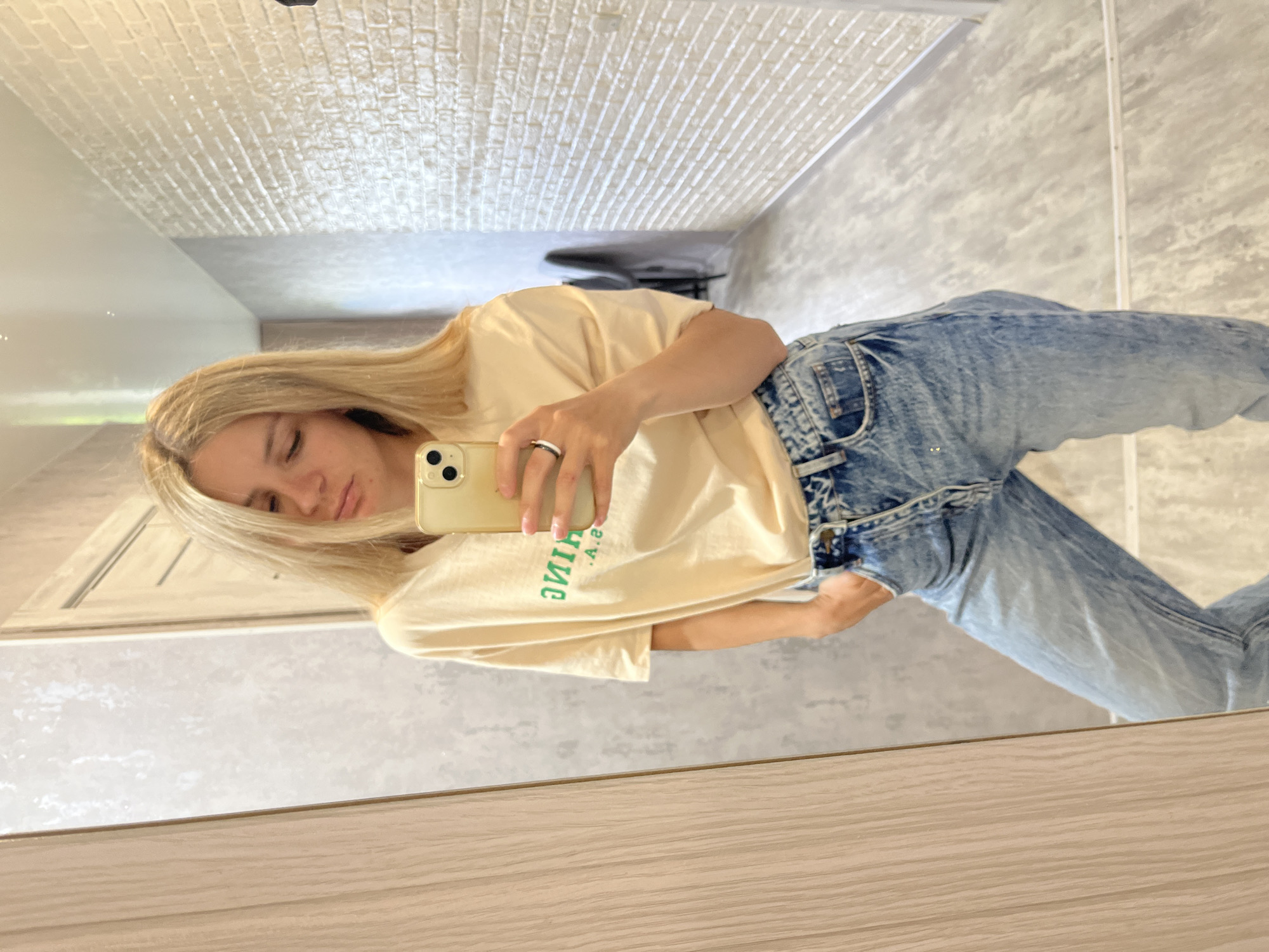 A woman with blonde hair is taking a mirror selfie in a room. She is wearing a light yellow t-shirt with green lettering and light blue distressed jeans. Her left hand is in her pocket. A smartphone is visible in her right hand, held up to take the photo. The background shows a hallway with a tiled wall and a wooden door. The scene is likely indoors, possibly a residential or hotel setting, in Chaikovsky, Russia.