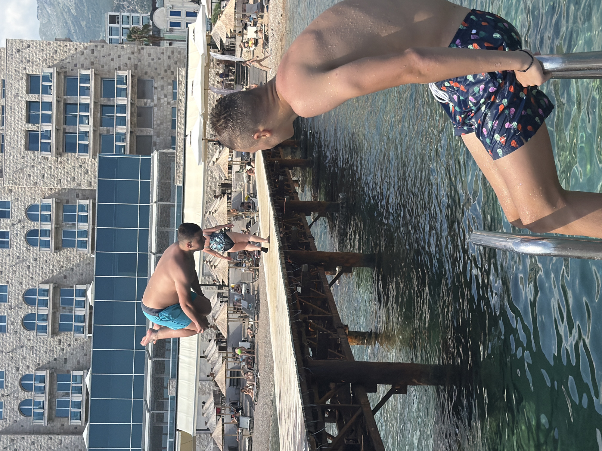 The image captures a lively scene at a beach resort in Budva, Montenegro. Two young individuals are the central focus. One person, wearing blue swim trunks, is captured mid-air as they are about to jump into the clear, turquoise water. Their body is contorted in a dynamic pose, suggesting the excitement of the jump. The other person, dressed in patterned swim trunks, is holding onto a metal railing and appears to be watching the jumper or preparing to follow.
The background reveals a bustling beach with numerous umbrellas providing shade to sunbathers and a large, modern building constructed with stone and glass. The building's architecture is distinct, with repetitive rectangular windows and some arched elements. The water is calm, with sunlight reflecting off its surface, creating dappled patterns. The overall atmosphere suggests a warm, sunny day at a popular tourist destination. The presence of beach umbrellas and people lounging indicates it's likely daytime, possibly late morning or afternoon. The image conveys a sense of youthful energy and recreational activity. No visible text is present in the image.