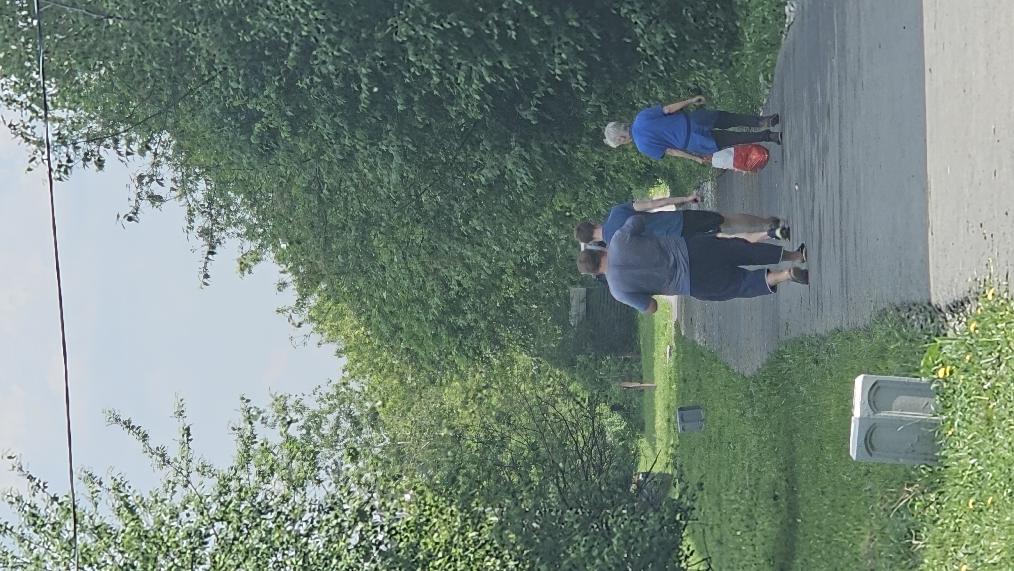 A group of three people and an elderly woman are walking down a concrete road on a sunny day. The road is flanked by lush green trees and grass on both sides. The elderly woman is carrying a red and white plastic bag. The location is the city of Unknown, Unknown.