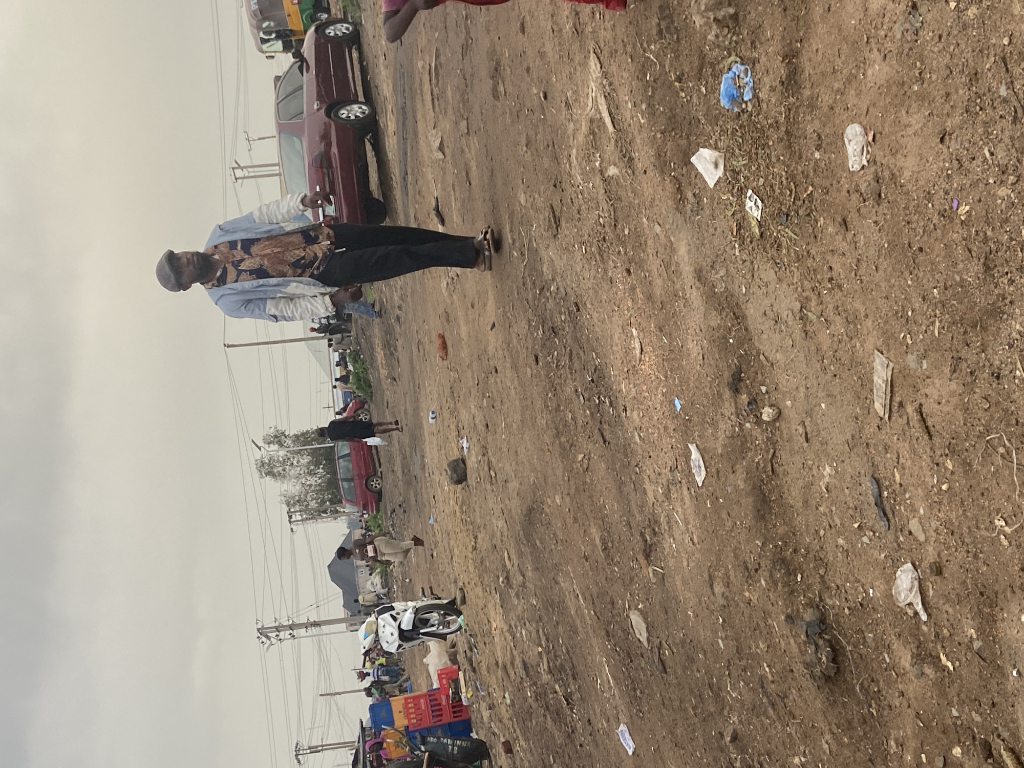 The image depicts a dusty, outdoor market scene in Bukuru, Nigeria. A man wearing a patterned shirt, a light blue jacket, and a cap is walking towards the right of the frame. He is wearing dark pants and sandals. In the background, a red sedan is parked, and other vehicles, including what appears to be a commercial tricycle (keke), are visible. Several individuals are present in the background, some appearing to be vendors or shoppers. A white motorcycle is parked in the middle left of the frame, next to stacks of red and blue crates. The ground is uneven and covered in dirt and scattered debris, including plastic bags and bottles. Overhead, a cloudy sky is visible with electrical wires strung across poles. The overall environment suggests a busy, potentially informal commercial area with a significant amount of litter.