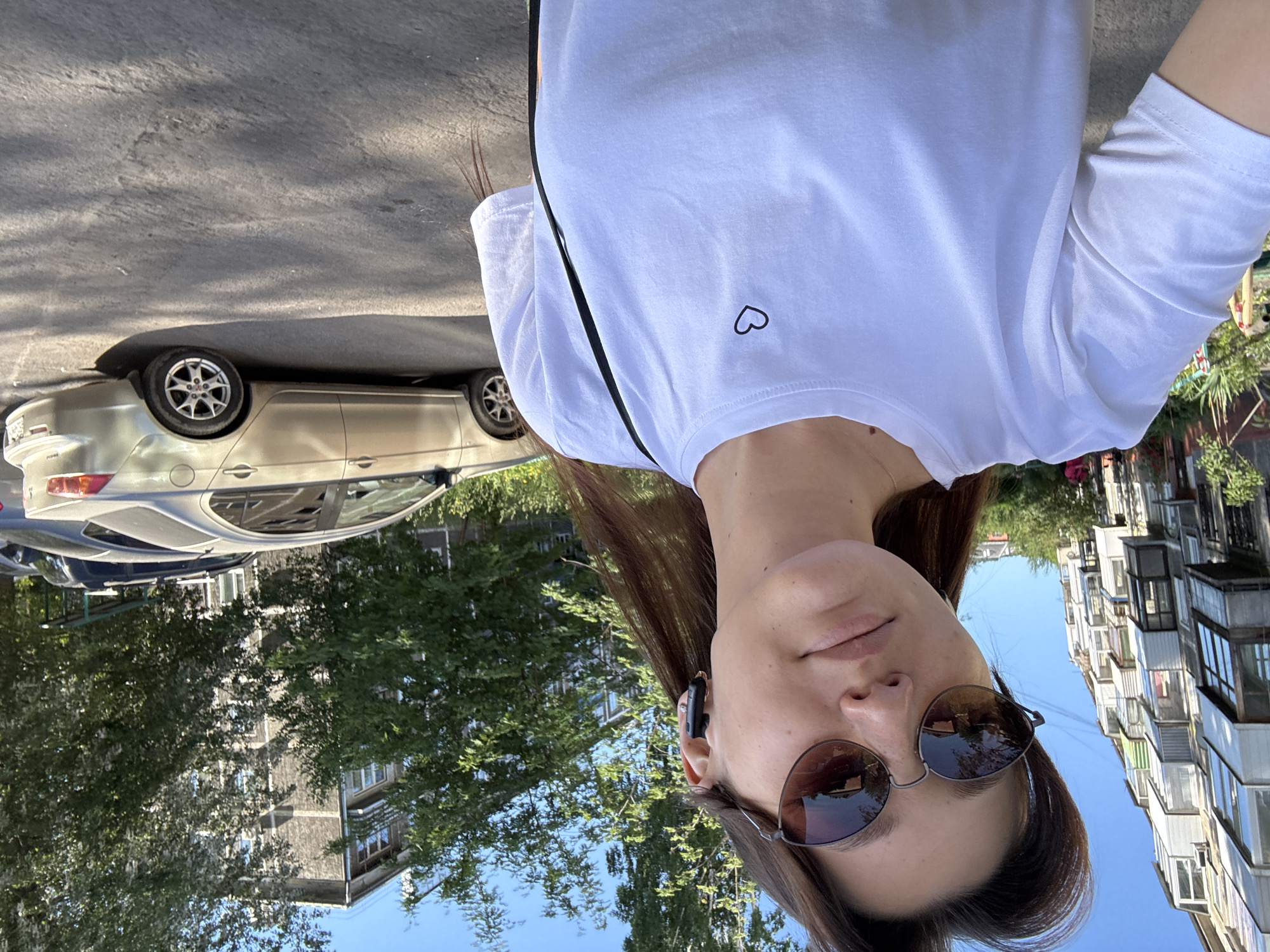 A young woman with brown hair, wearing sunglasses and a white t-shirt with a small black heart emblem, is taking a selfie outdoors in a city. She is also wearing black earbuds. Behind her, a light-colored sedan is parked on a paved surface. In the background, there are apartment buildings and trees. The scene suggests a sunny day in an urban environment.