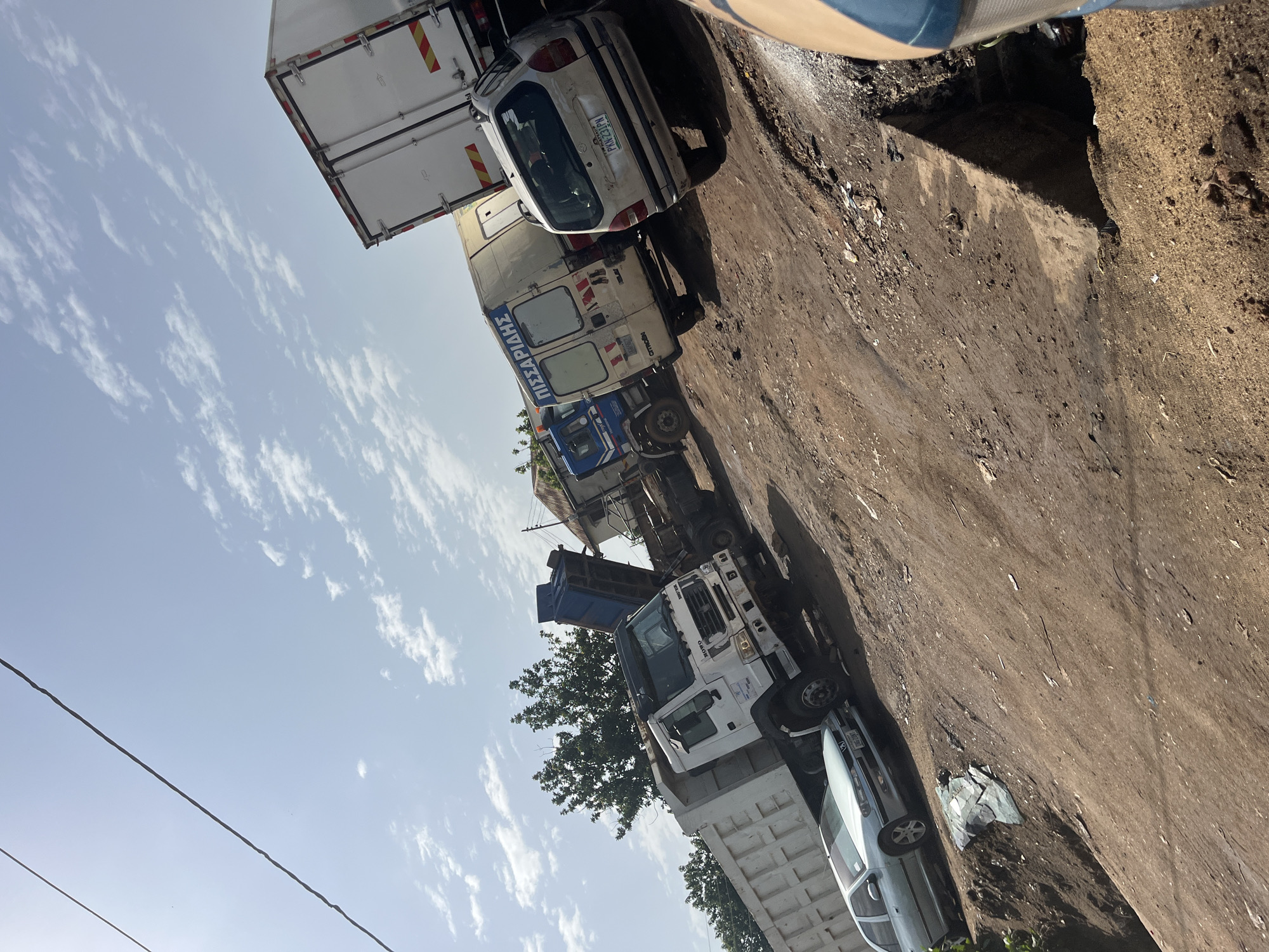 In Bukuru, Nigeria, a low-angle, sunlit outdoor scene is dominated by several large trucks and a car parked on a dirt lot. A white dump truck with its bed partially raised is in the foreground, to the left, next to a light-colored sedan. Behind the dump truck, a white box truck with writing in a foreign language is visible, followed by a blue and white truck. Further back, a white semi-trailer truck with reflective markings is parked. The ground is uneven and covered with dirt, gravel, and debris. Trees are visible in the background, and a building with a roof is to the right. The sky is blue with scattered white clouds, and power lines run diagonally across the upper left portion of the frame.