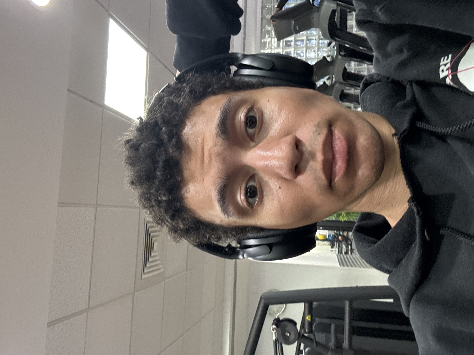 The image depicts a person with curly dark hair wearing black headphones and a black hooded sweatshirt. The person is indoors, likely in a gymnasium or fitness center, based on the presence of exercise equipment visible in the background. The setting features tiled walls and fluorescent lighting. The location in London, United Kingdom cannot be determined from the image.