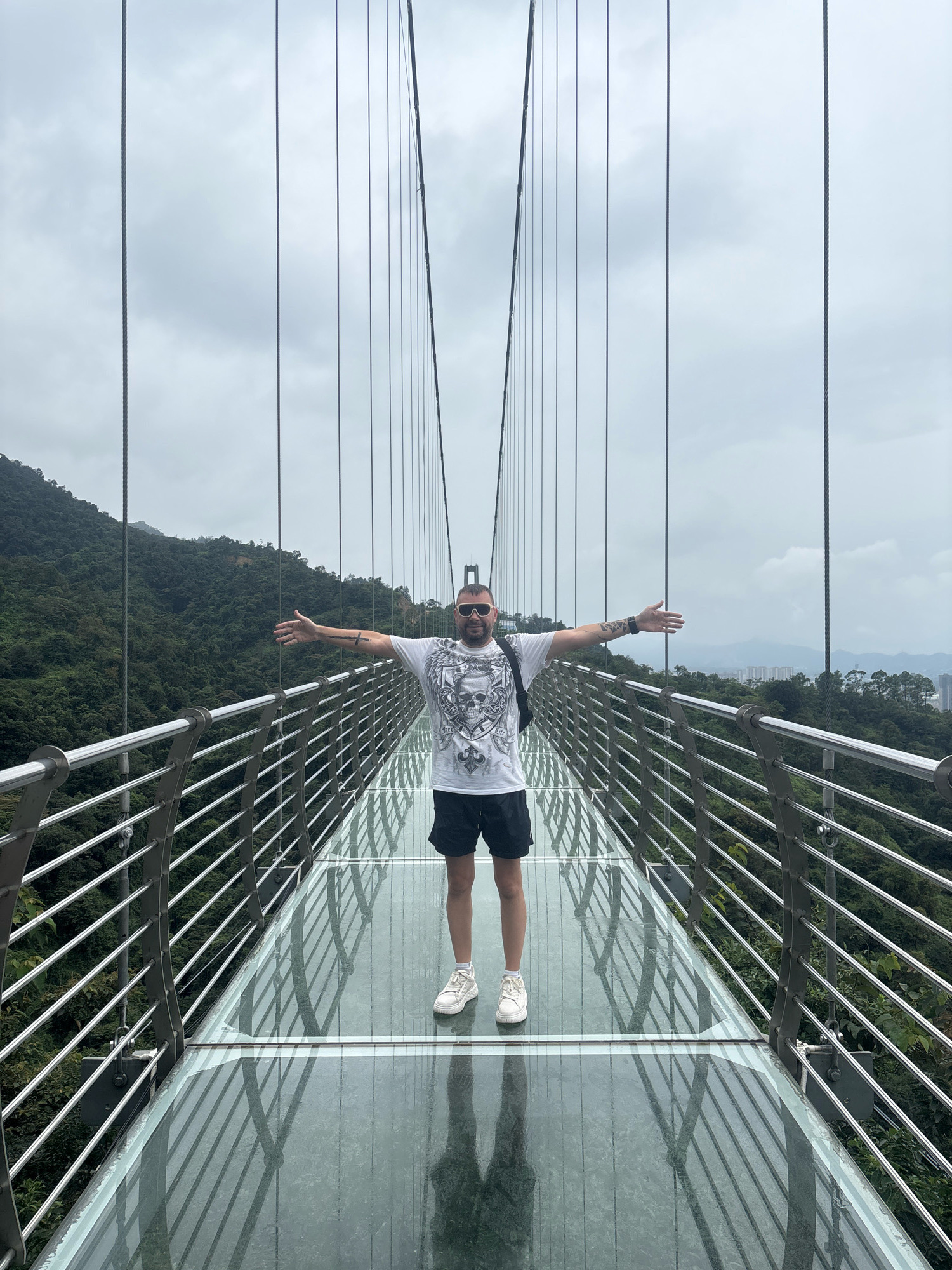 A man stands with his arms outstretched on a glass-bottomed suspension bridge. He is wearing a white t-shirt with a graphic print, black shorts, white sneakers, sunglasses, and a backpack. The bridge is suspended over a lush, green, mountainous landscape. In the distance, a cityscape is visible under a cloudy sky. The bridge's glass floor reflects the cables supporting the structure and the surrounding scenery. This bridge is located in Foshan, China, specifically at the Nanhai Xiqiao Mountain Glass Bridge.
