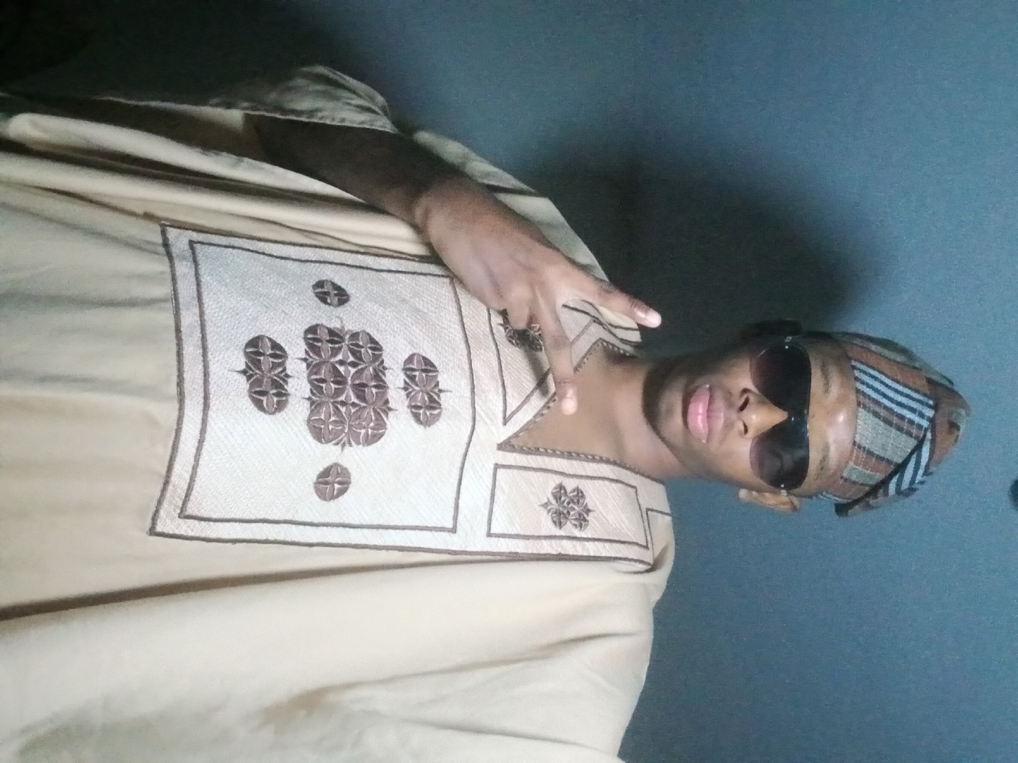 A young person wearing a light-colored, embroidered traditional garment and a patterned headwrap is shown from the chest up, against a plain, gray background. The person is wearing dark sunglasses and has their right hand raised, forming a peace sign. The garment has intricate brown embroidery in a geometric pattern on the chest area. The lighting in the scene is dim, with a shadow cast on the left side of the background.