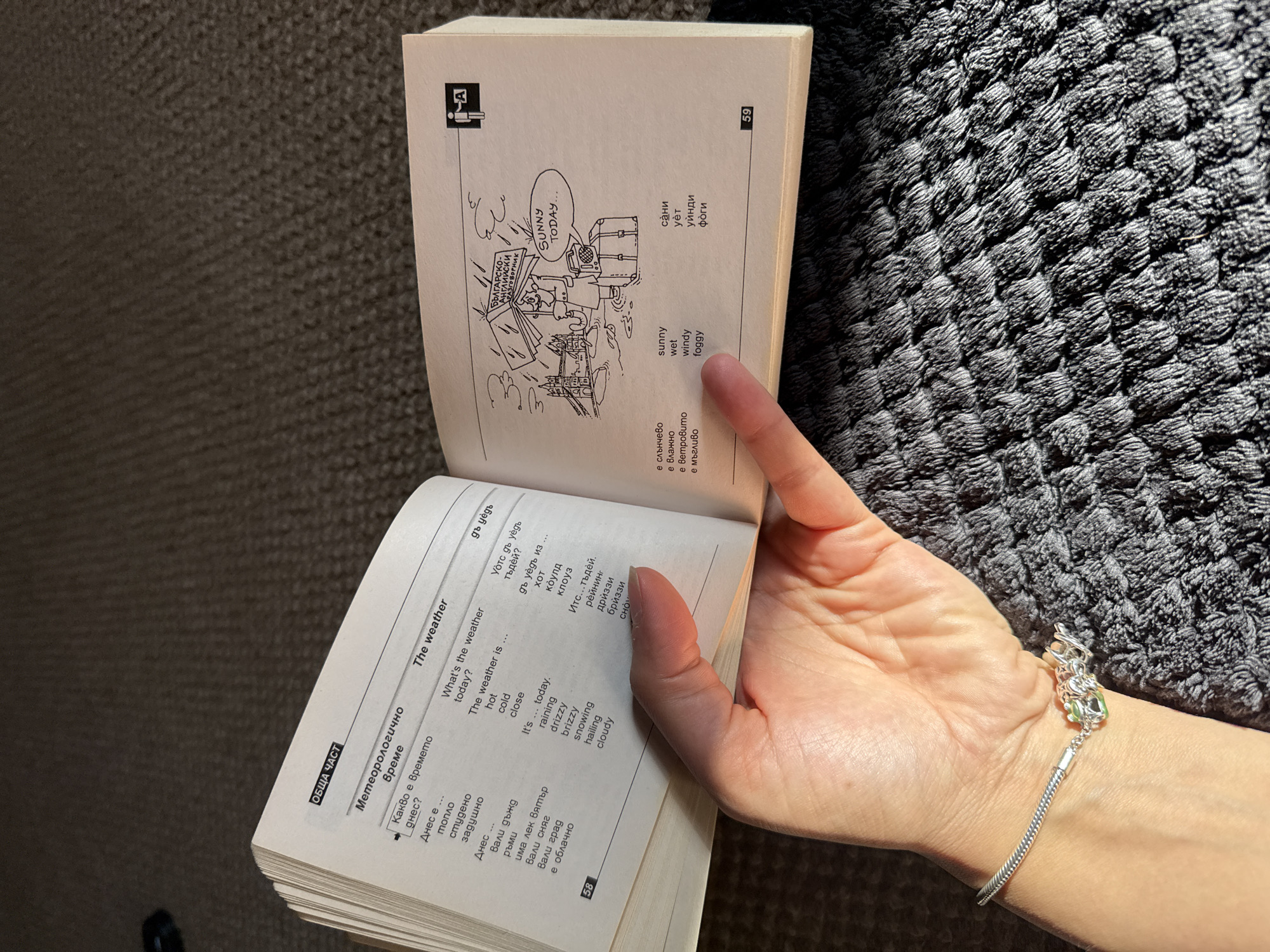 The image shows an open book with the title "The Weather" in both English and Bulgarian. The pages contain vocabulary and phrases related to weather conditions. On one page, there is a cartoon depicting a person with luggage in a sunny setting, labeled "Sunny Today..." and a drawing of a famous Great Yarmouth landmark. The book is being held open by a hand wearing a silver bracelet with decorative charms. The background is a textured gray fabric.