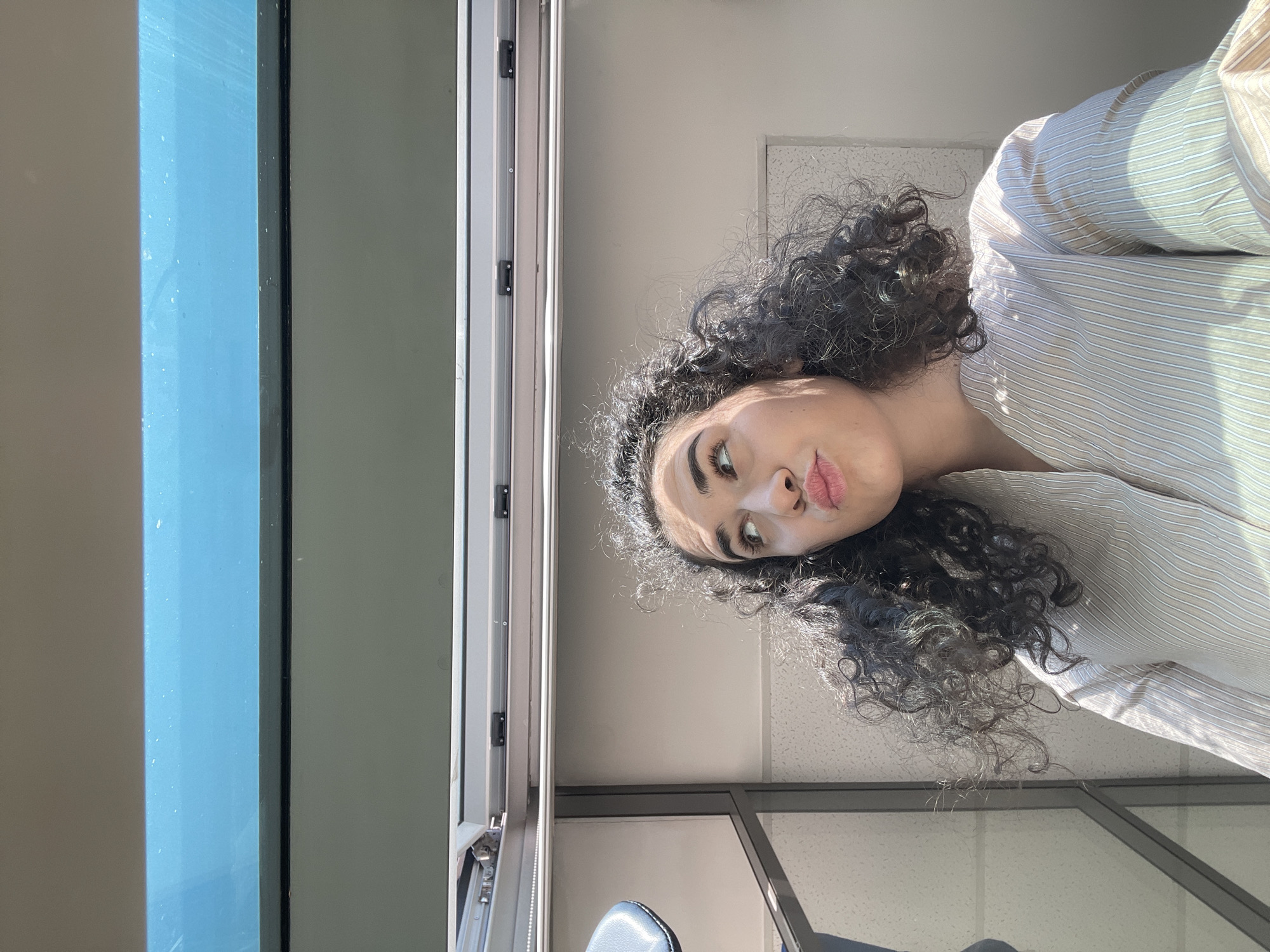 An overcast sky with some white clouds is visible through a large, angled window. Below the window, a woman with dark, curly hair is looking upwards and to her right with her lips puckered. She is wearing a light-colored, pinstriped shirt. The environment appears to be an indoor space with a white textured wall and metallic framing visible. The lighting suggests it is daytime, with sunlight casting shadows and highlighting the woman's hair. The location is an indoor space with a window, possibly in Sofia, Bulgaria, but no specific landmarks are visible to confirm this.