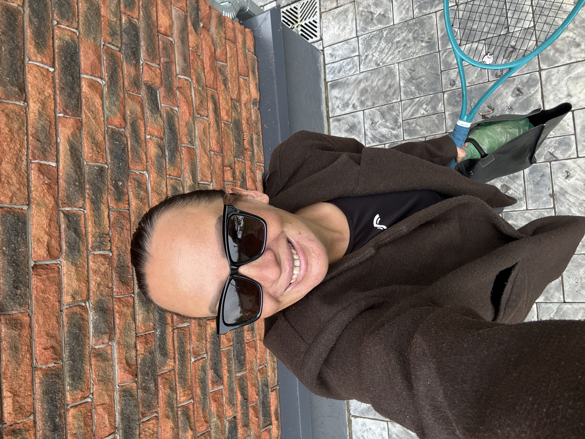 A selfie-style, medium-angle shot presents a person holding a tennis racket and bag, with a brick wall to the left and a tiled surface to the right. The person is wearing a dark-colored jacket over a black shirt with a white logo, and large black sunglasses. They are smiling with visible teeth. The tennis racket is light blue with a blue grip, and a black bag sits near it, seemingly holding other items. The wall appears to be made of orange bricks with grey mortar, and the tiles on the ground are grey with a textured surface. A metal drain is visible near the wall. The photograph is rotated counterclockwise. No information suggests it was taken in Chaikovsky, Russia.