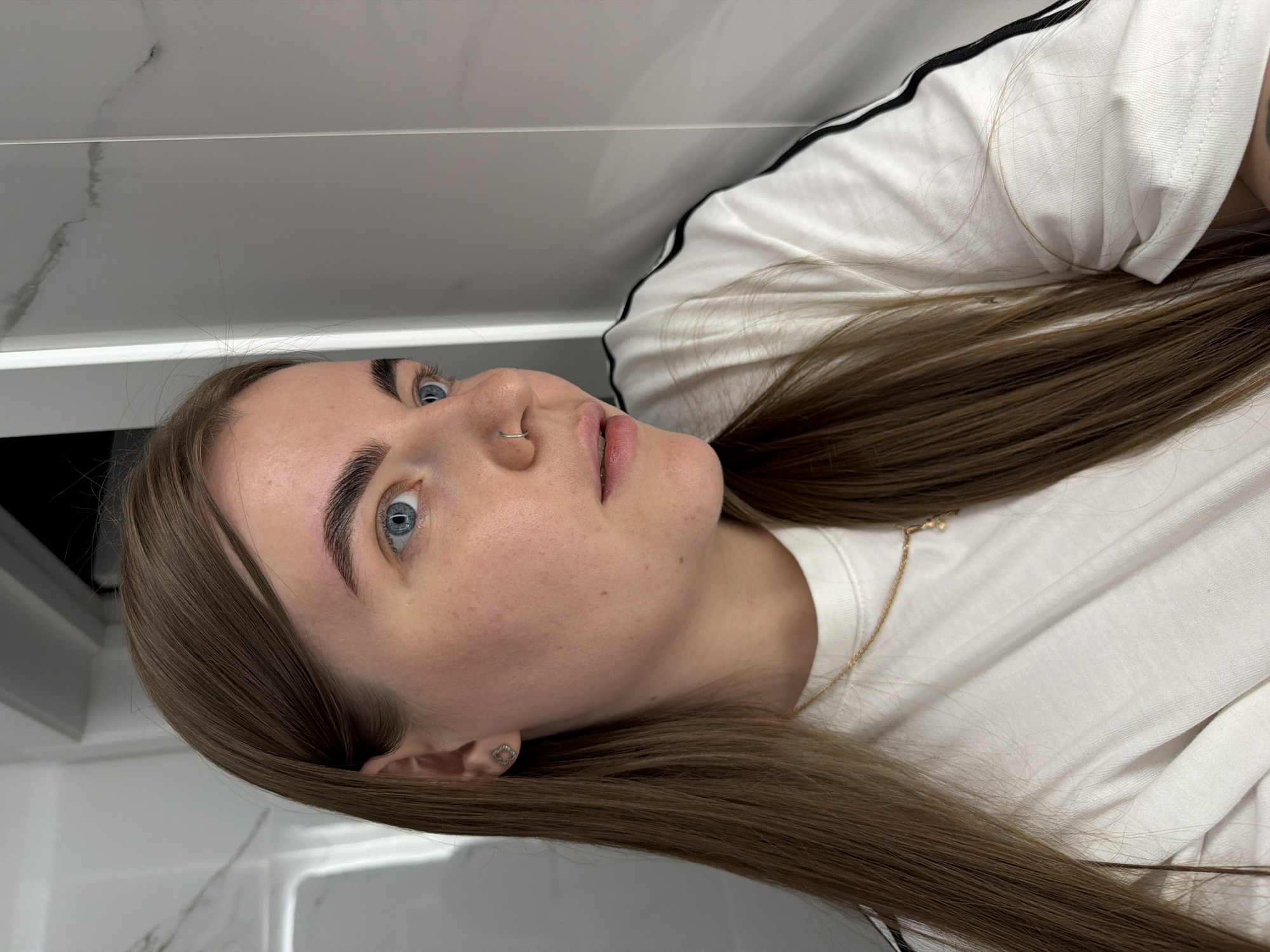 A young woman with light brown hair and blue eyes is seen from a low angle. She is lying down against a white and light gray background, likely a wall or counter. She has a nose ring in her left nostril and a small stud earring. Her eyebrows are well-defined. She's wearing a white t-shirt with black stripes on the sleeve. A delicate gold necklace with a pendant is visible. There is no indication of the location being in Chaikovsky, Russia.