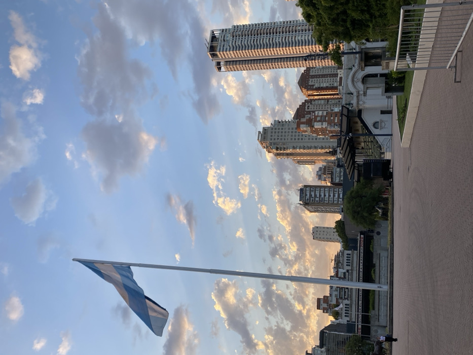 The image depicts an urban outdoor scene at sunset or sunrise, located in the Plaza República de Perú / Plaza Naciones Unidas area, at the boundary of Palermo Chico and Recoleta, Buenos Aires, Argentina.
In the upper left quadrant, a large Argentine flag, featuring light blue, white, and light blue horizontal stripes, flies partially unfurled from a tall, light-colored flagpole against a light blue sky punctuated by numerous altocumulus clouds, which are illuminated with warm, golden hues from the low sun.
The background features a skyline of modern high-rise residential buildings, including the prominent, slender Le Parc Figueroa Alcorta tower on the far right, all catching the golden light. Below these towers and to the left, a large, ornate white classical-style building, identified as the Palacio de Exposiciones (Exposition Palace) due to its architecture and visible text "CULTIVAR EL SUELO SERVIR A LA PATRIA 2023" on a lower section, forms part of the mid-ground. In front of this building, a dark outdoor stage structure with visible stairs is present. To its left, a group of sculptures, including what appear to be horse figures, are visible on a slightly lower level.
The foreground consists of a wide, reddish-brown paved plaza. On the far left of this plaza, a single individual wearing dark clothing walks towards the right side of the frame. To the immediate right in the foreground, a silver-colored metal crowd barrier is partially visible, and a section of green lawn with dense foliage from trees borders the paved area.